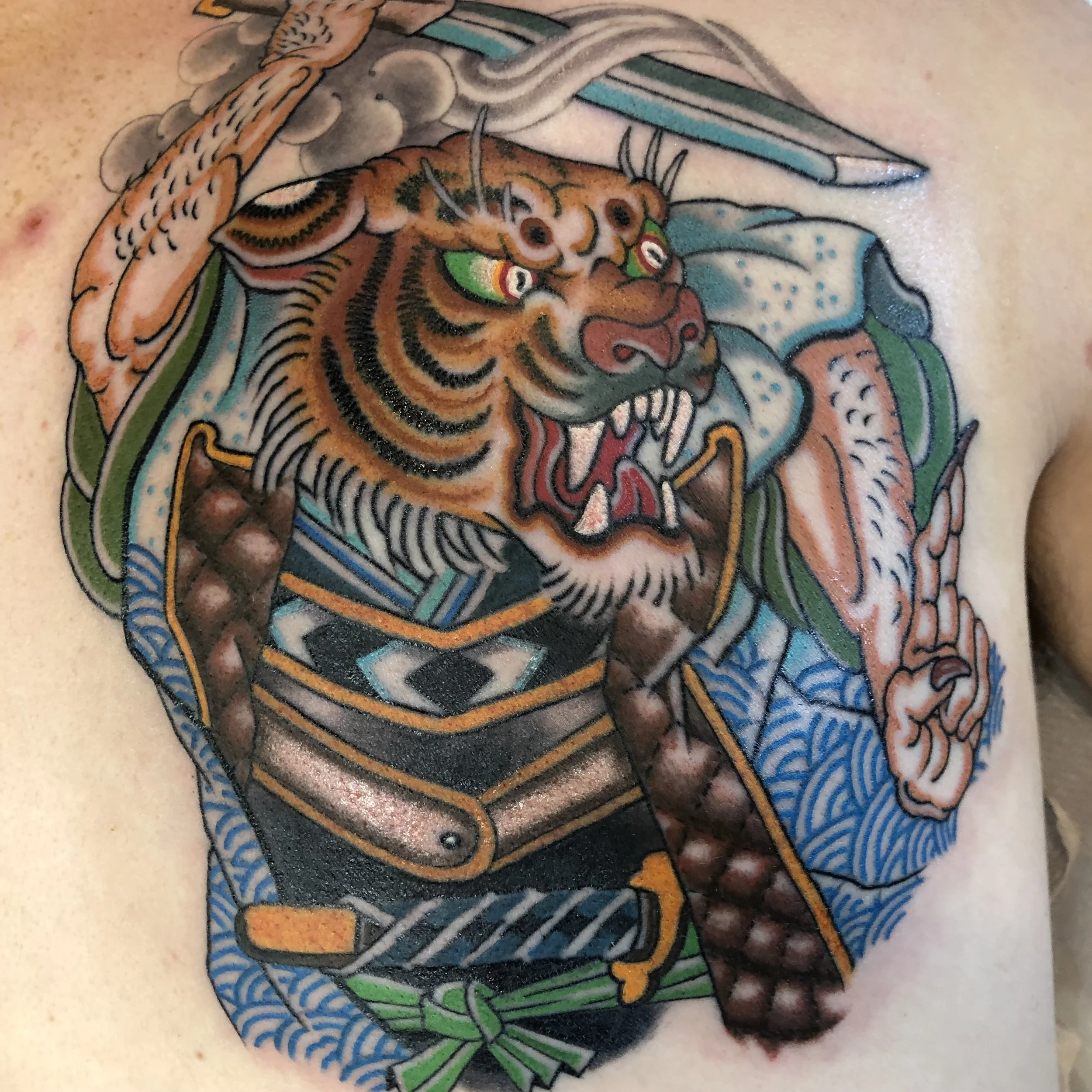Tattoo of a fierce tiger with green eyes, bared teeth, and detailed orange, black, and white fur, wearing a traditional Japanese armor, on skin with a background of clouds and waves.