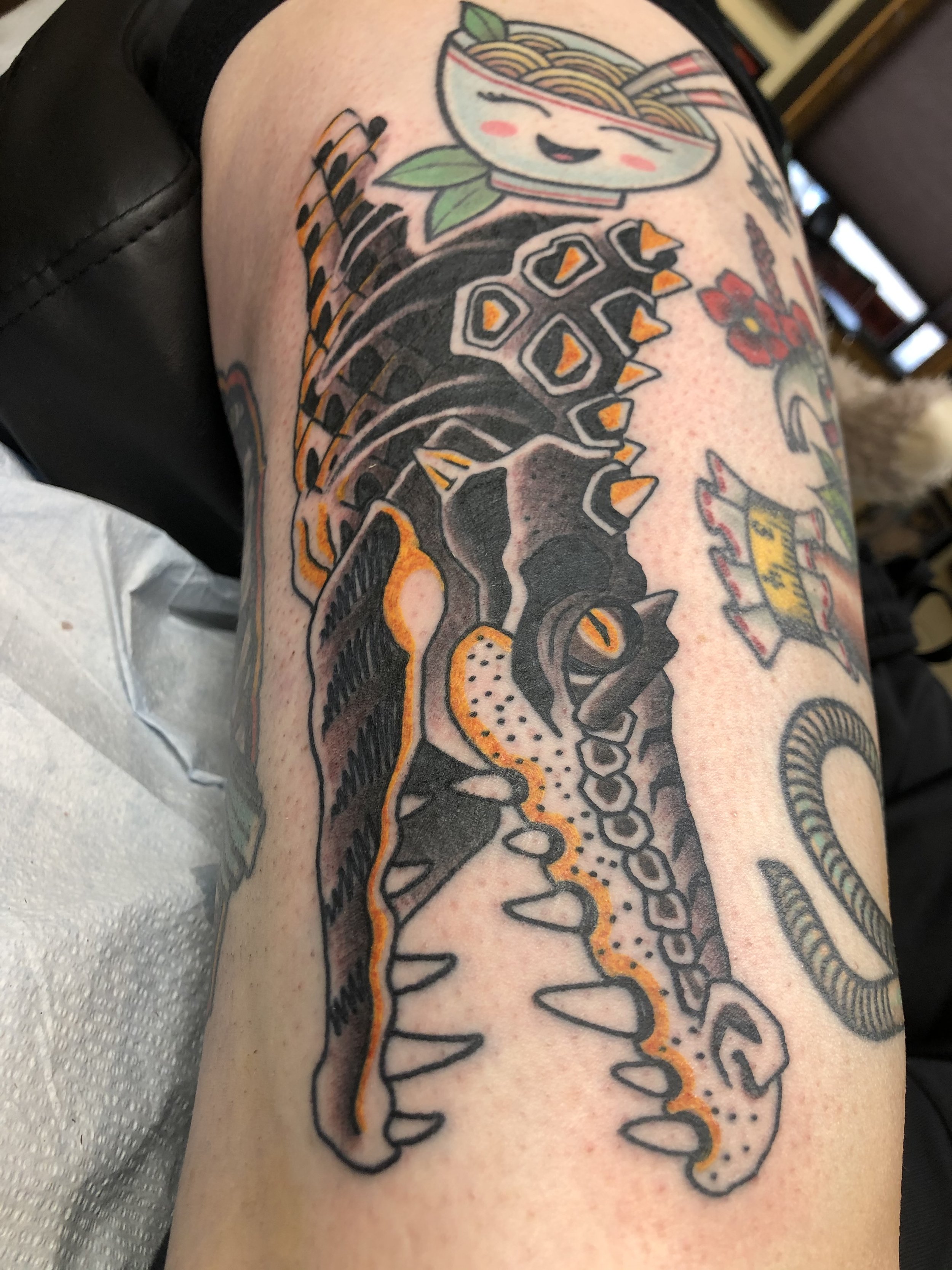 Colorful tattoo of an alligator with an orange outline and black details on a person's leg.