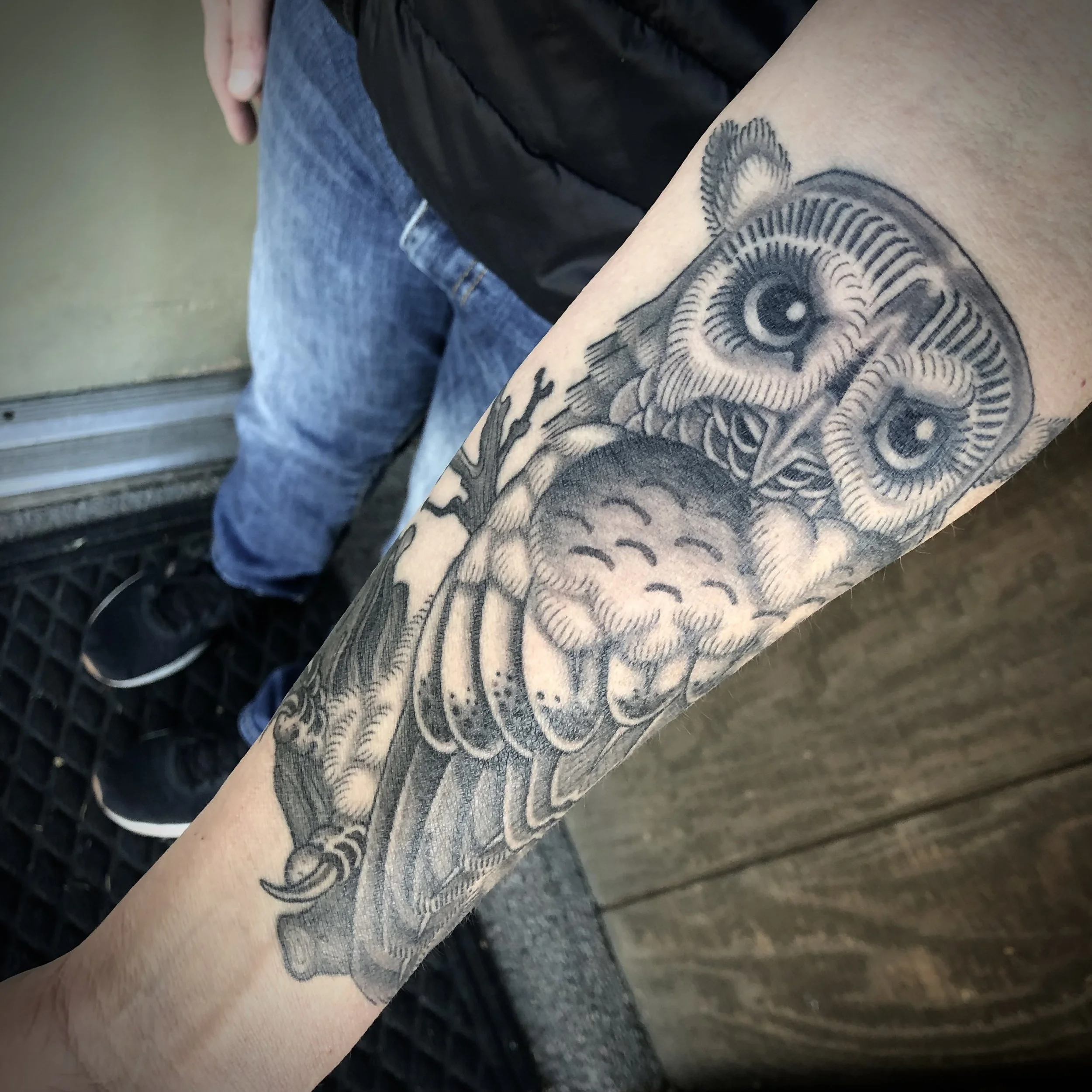 Black and gray tattoo of an owl on a person's forearm, featuring detailed feathers and large eyes.