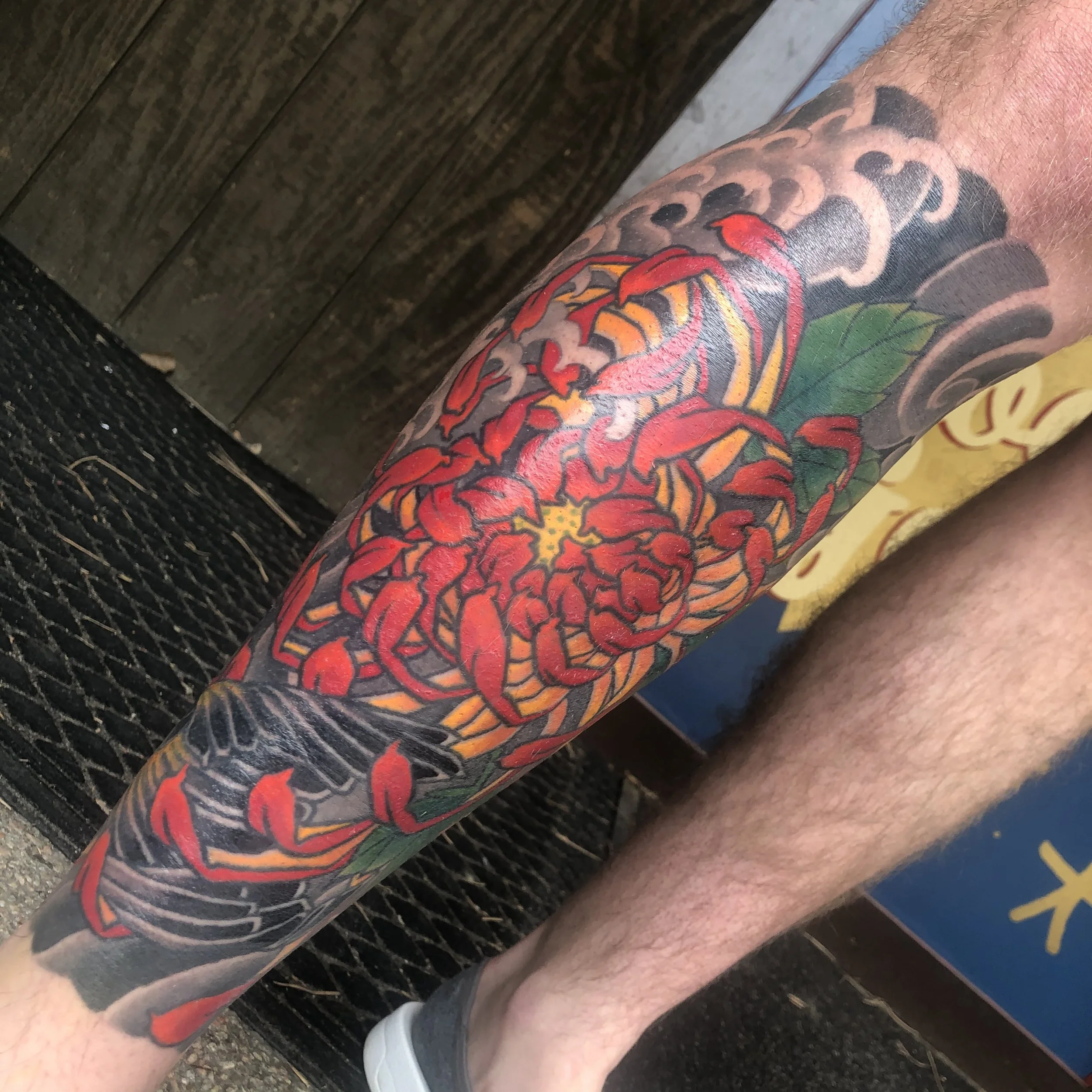 Colorful tattoo of a red and yellow chrysanthemum flower with green leaves on a person's leg.