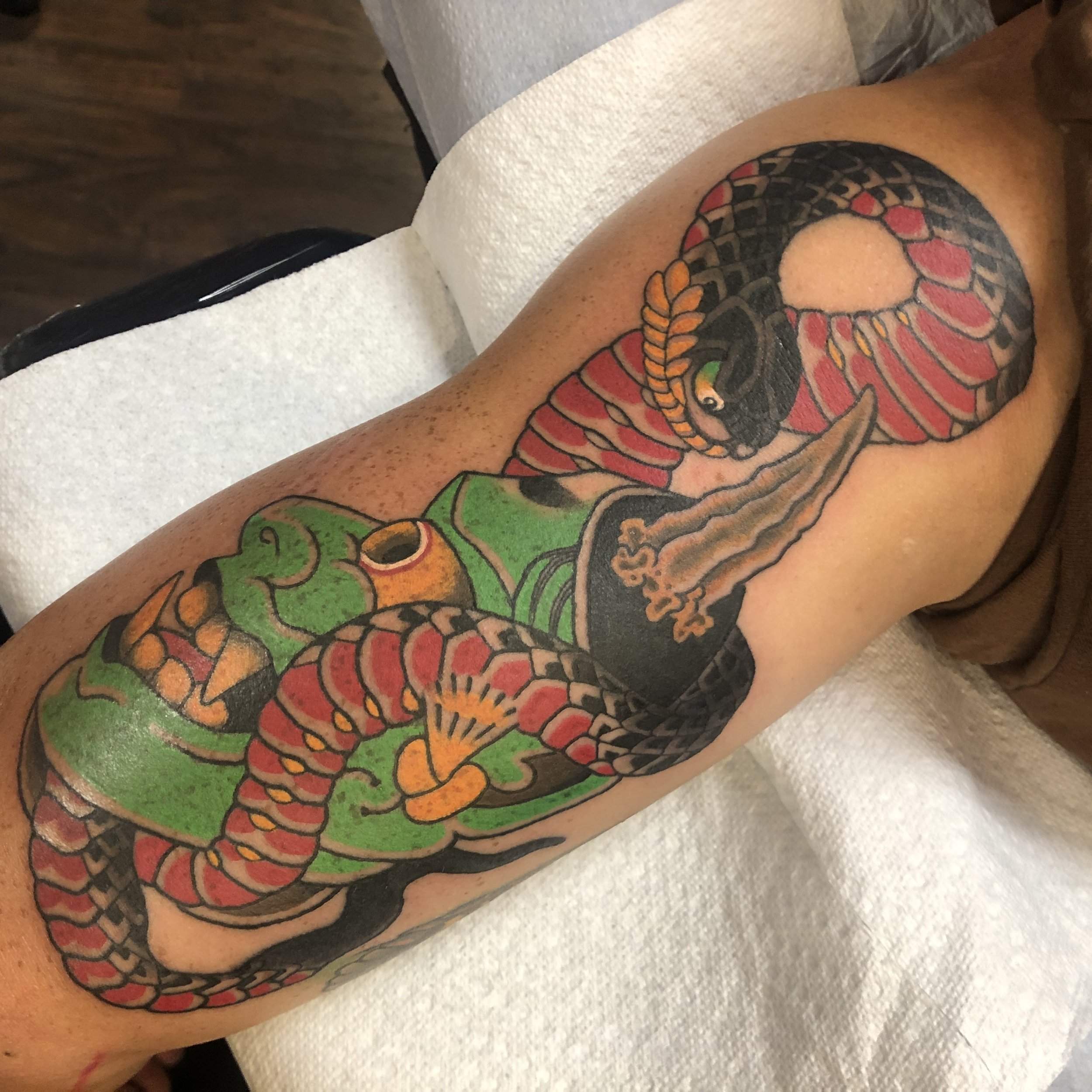 Colorful traditional-style snake and hannya tattoo on a person's upper arm, featuring a black and red snake intertwined with green and orange accents.
