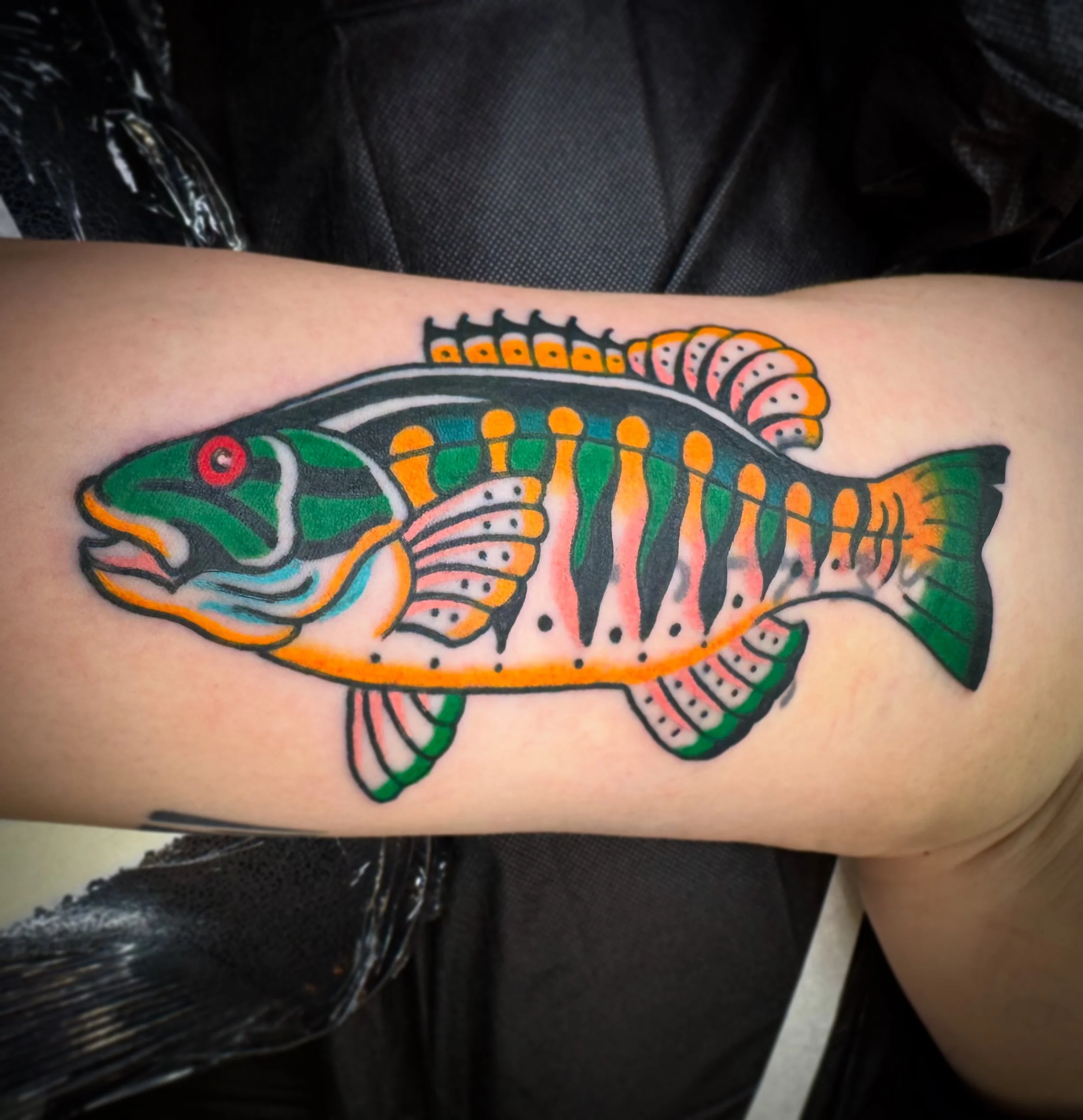 A colorful fish tattoo on a person's arm, featuring vibrant green, orange, pink, and black colors with detailed fins and stripes.