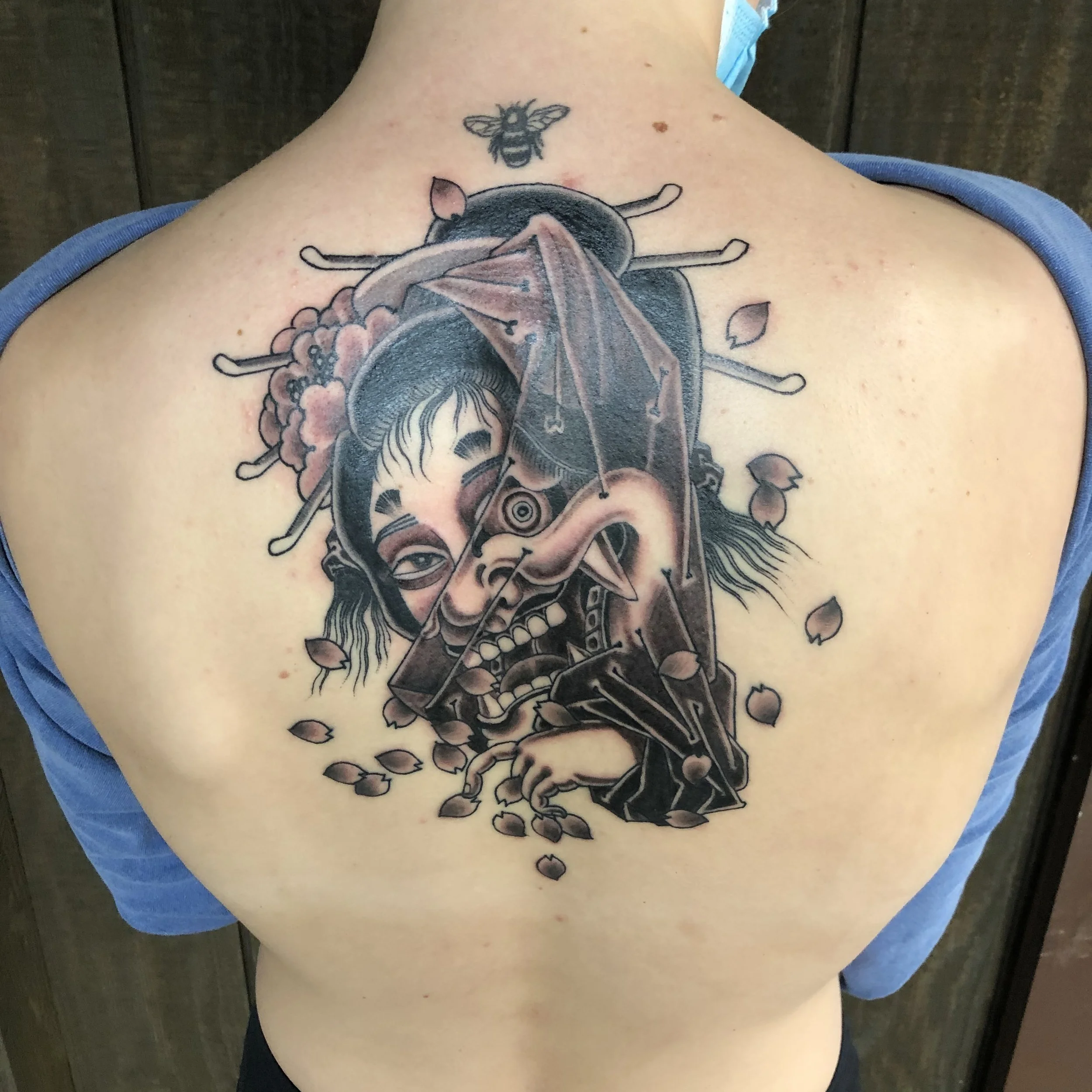 Tattoo of a Japanese geisha, partly hidden by a veil revealing a sinister demon with long hair, partly hidden , surrounded by falling blossom pedals on the person's back.