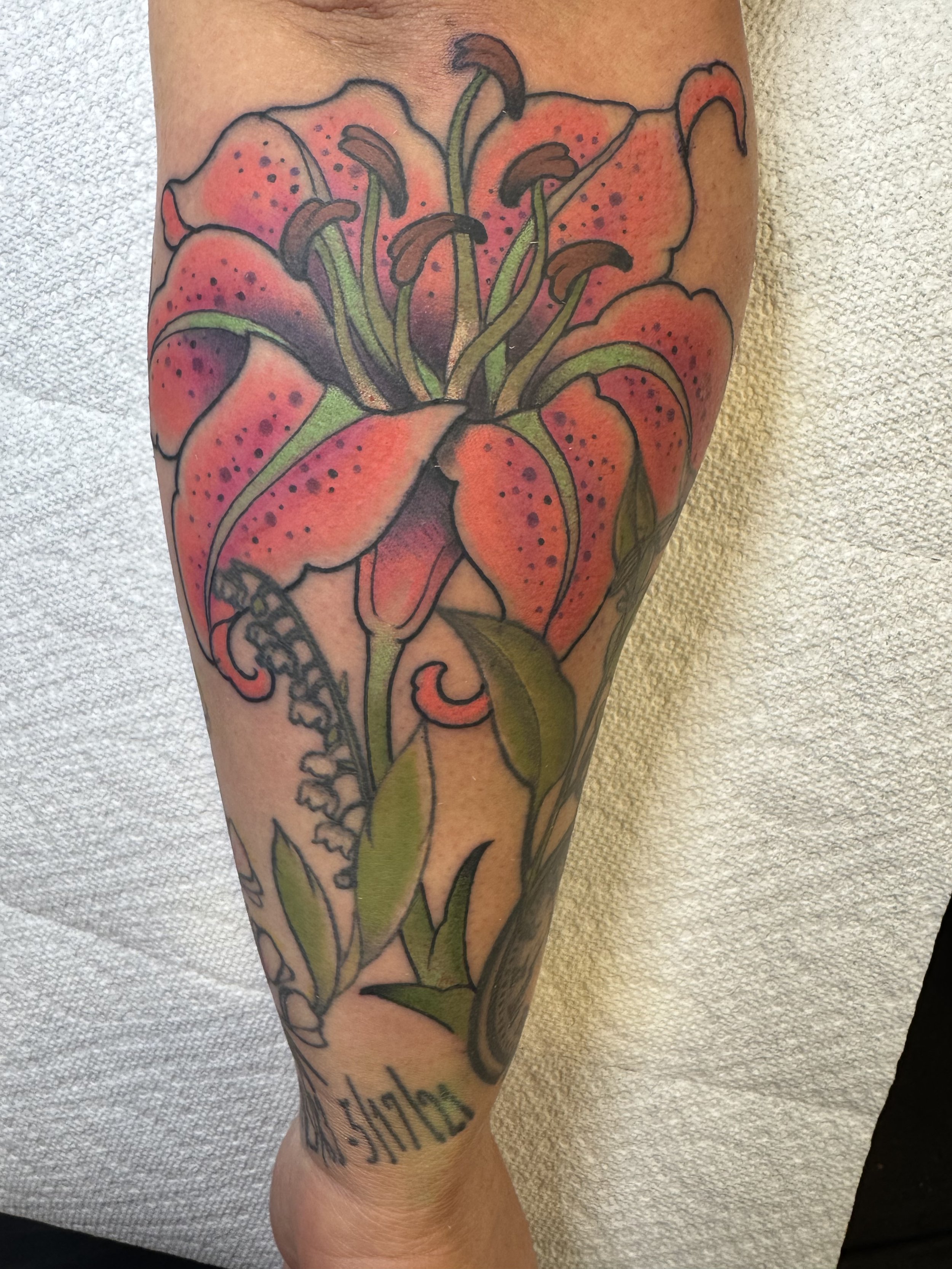 Colorful tattoo of a large pink lily with green leaves on a person's leg.