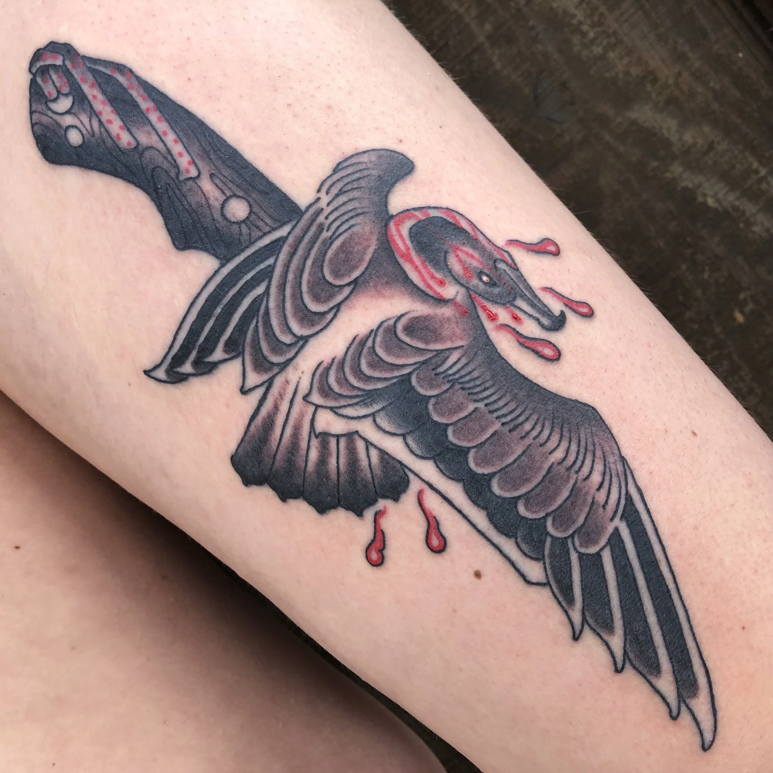 A tattoo of a bird and dagger with blood dripping from its beak and wounds, depicted in a traditional style with black and red ink.