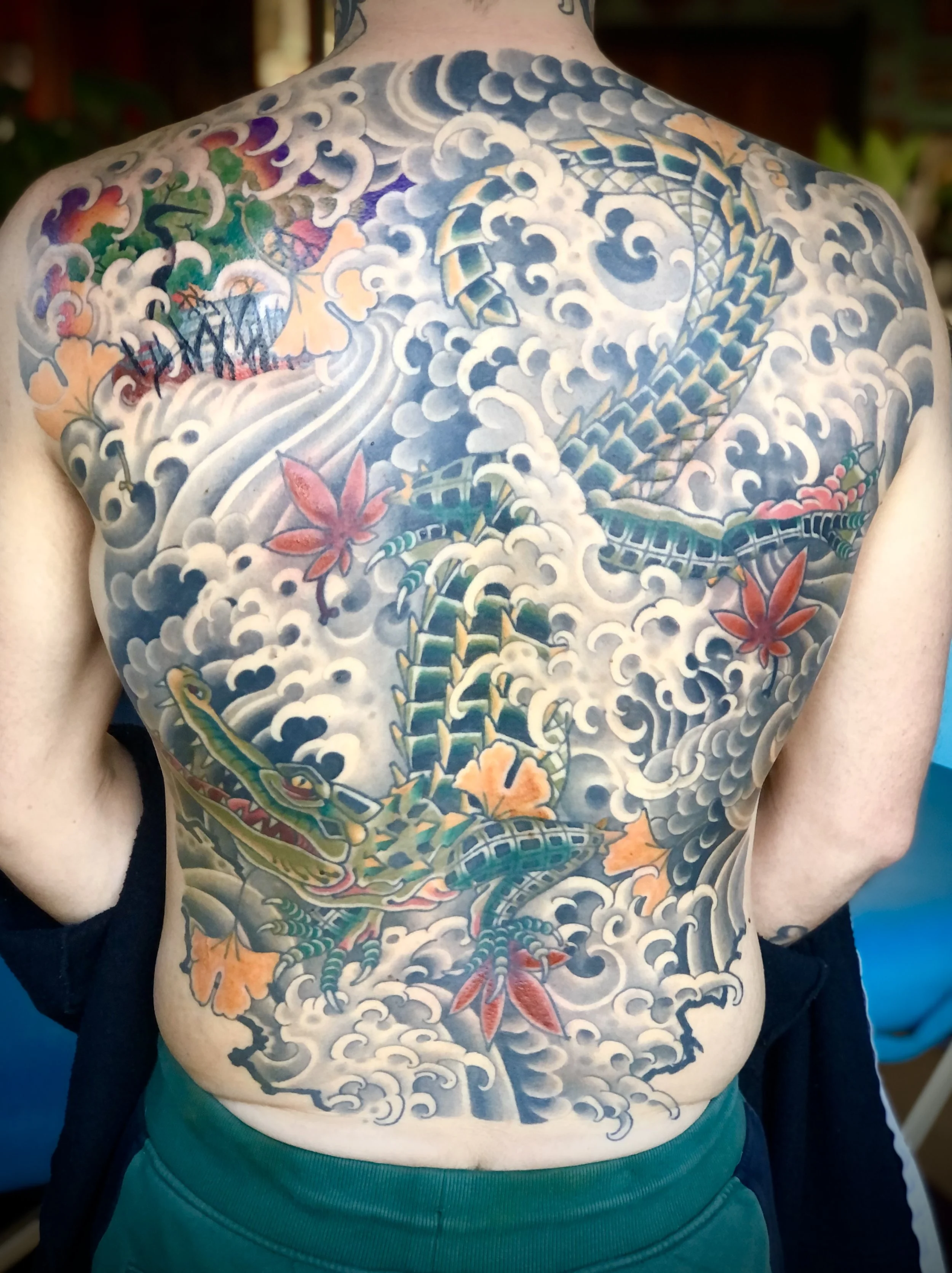 Colorful Japanese-style tattoo on a person's back featuring an alligator intertwined with clouds, red maple leaves, and a small landscape with trees and water.