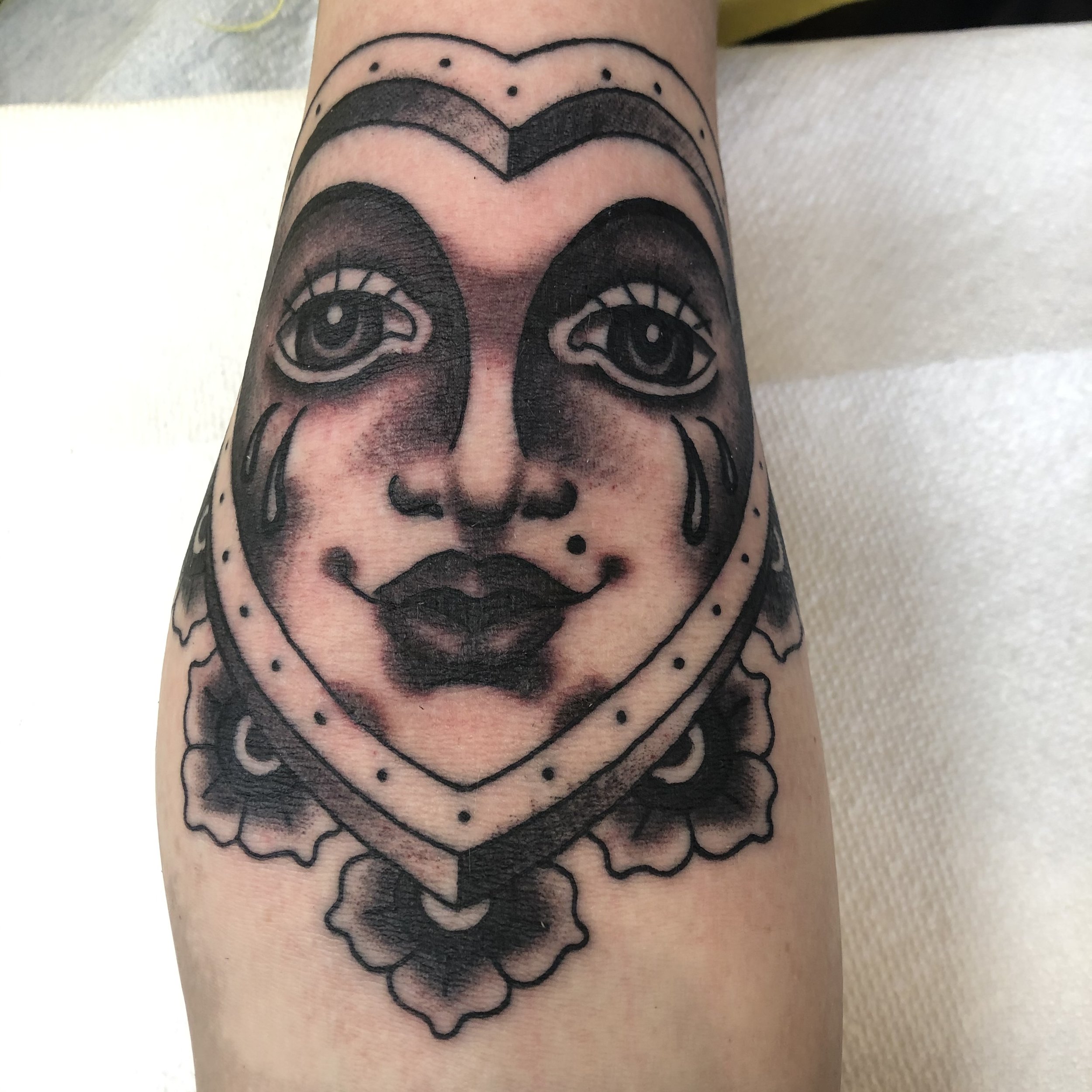 Black and gray tattoo of a woman's face with tears and lipstick inside a heart-shaped frame, with floral details.
