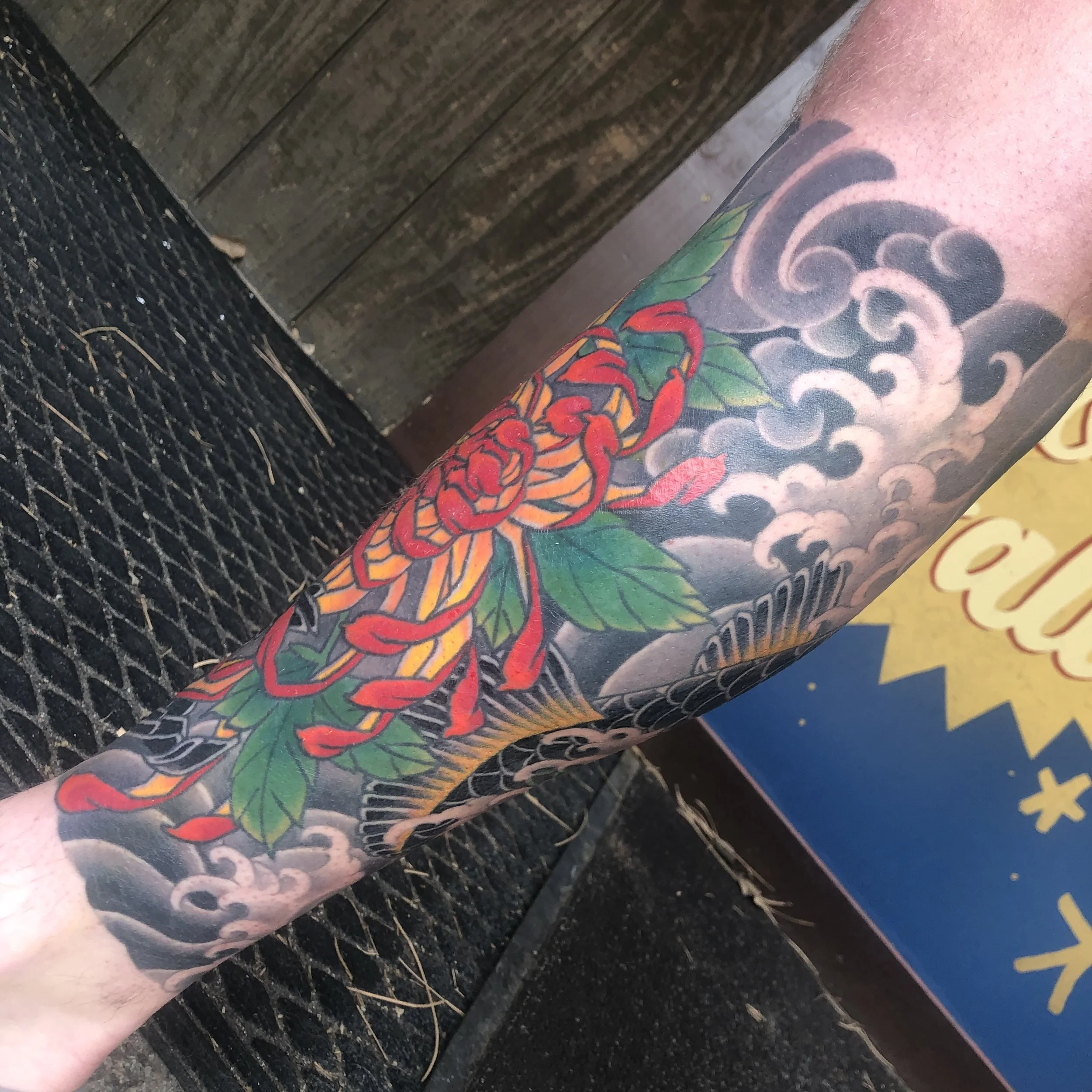 Colorful Japanese-style tattoo of a red chrysanthemum flower with green leaves, waves, and clouds on a person's leg.