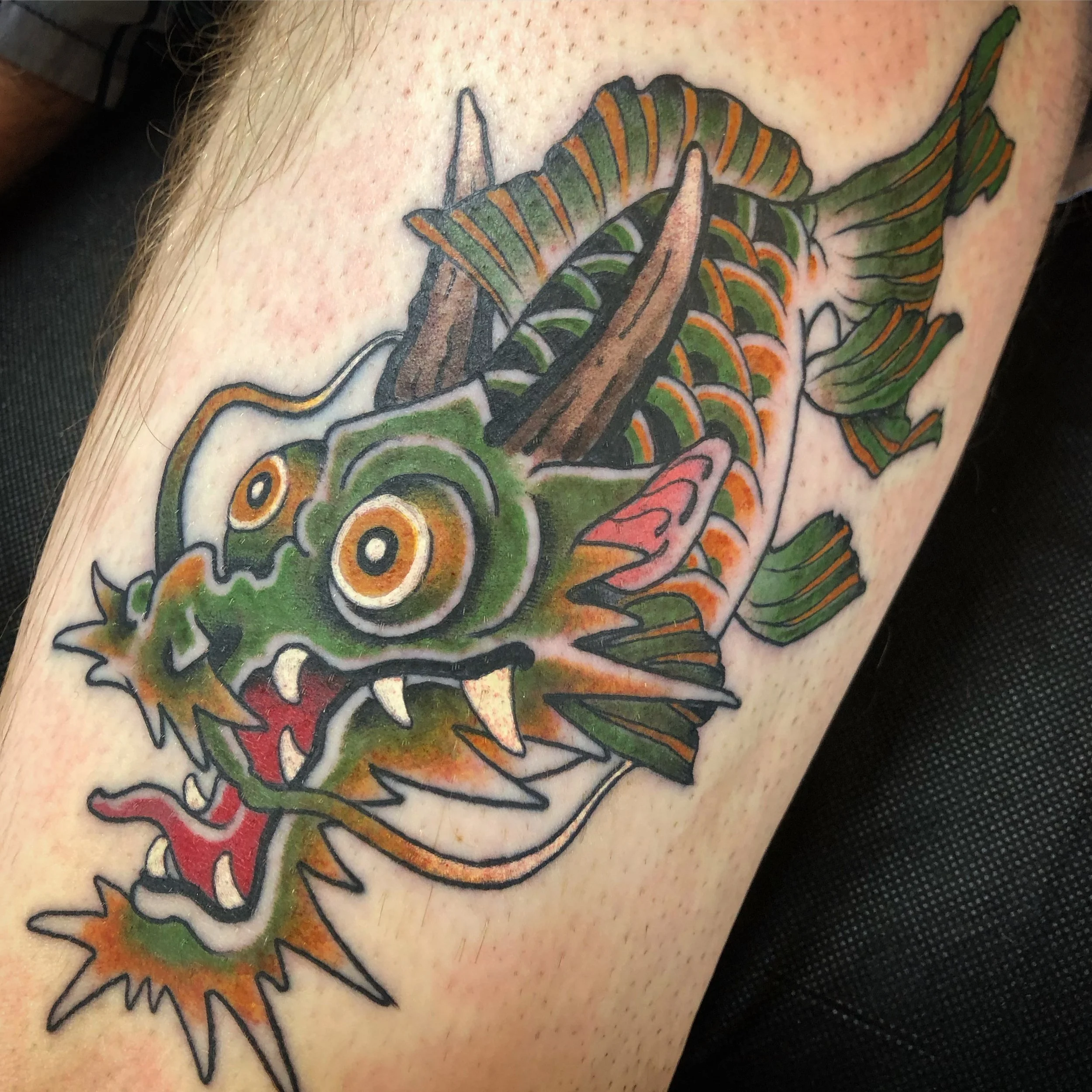 Colorful tattoo of a cartoon-like dragon fish with big eyes, sharp teeth, and spiky fins, in green, orange, and brown hues.