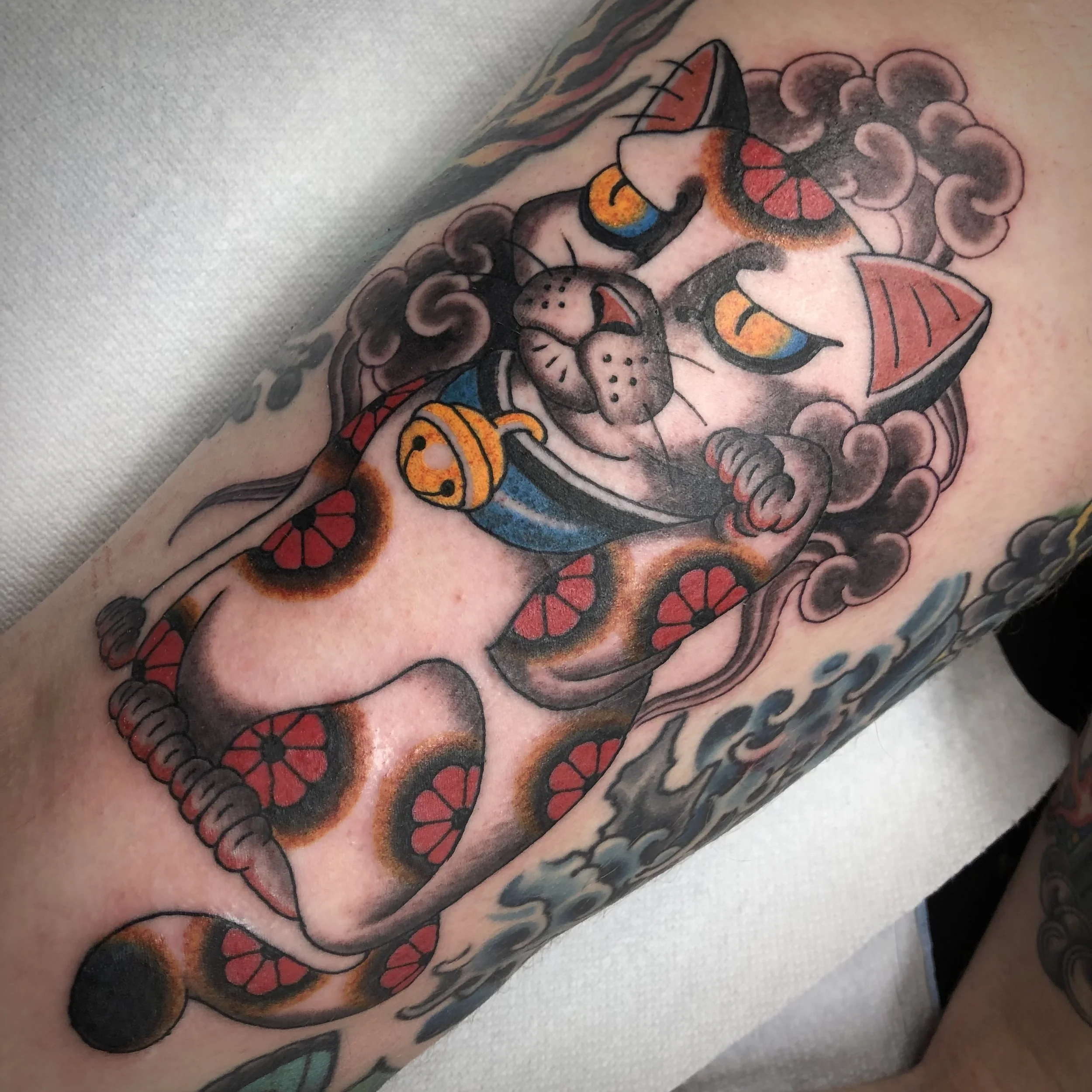 Traditional style tattoo of a beckoning cat with flowers and clouds.