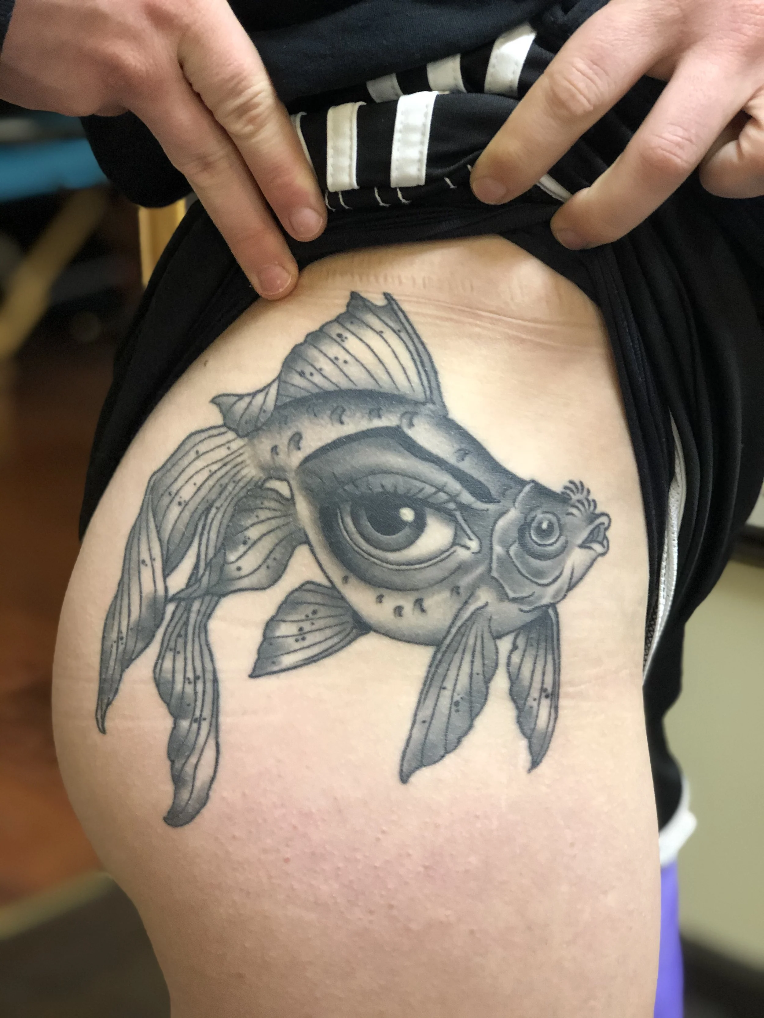 Tattoo of a fish with an eye as the main focus, detailed fins, and scales, on a person's upper thigh.