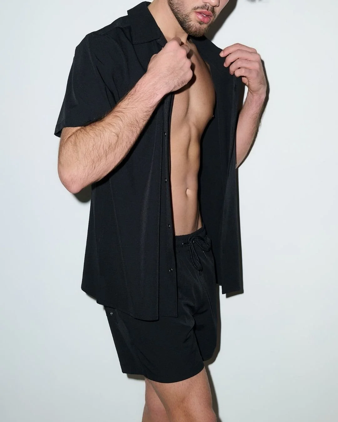 A man in a black shirt and shorts is partially unbuttoning his shirt to reveal his toned abdomen.