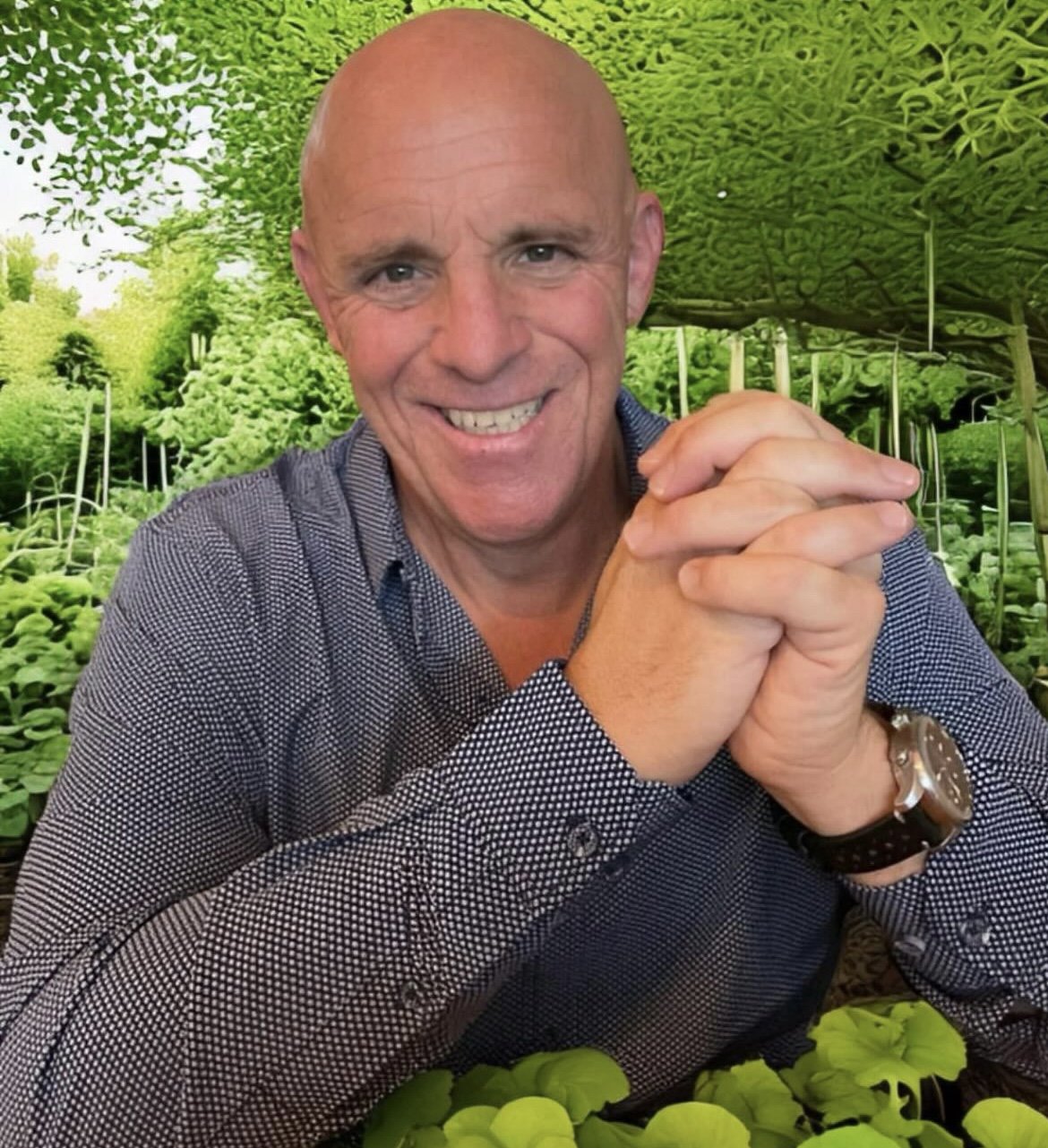 A man with a shaved head smiling and clasping his hands together, sitting in a lush green garden with various plants and trees.