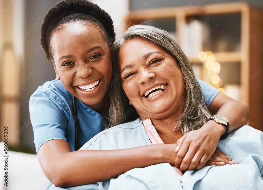 Young female healthcare worker hugging an older woman, both smiling warmly in a cozy room.