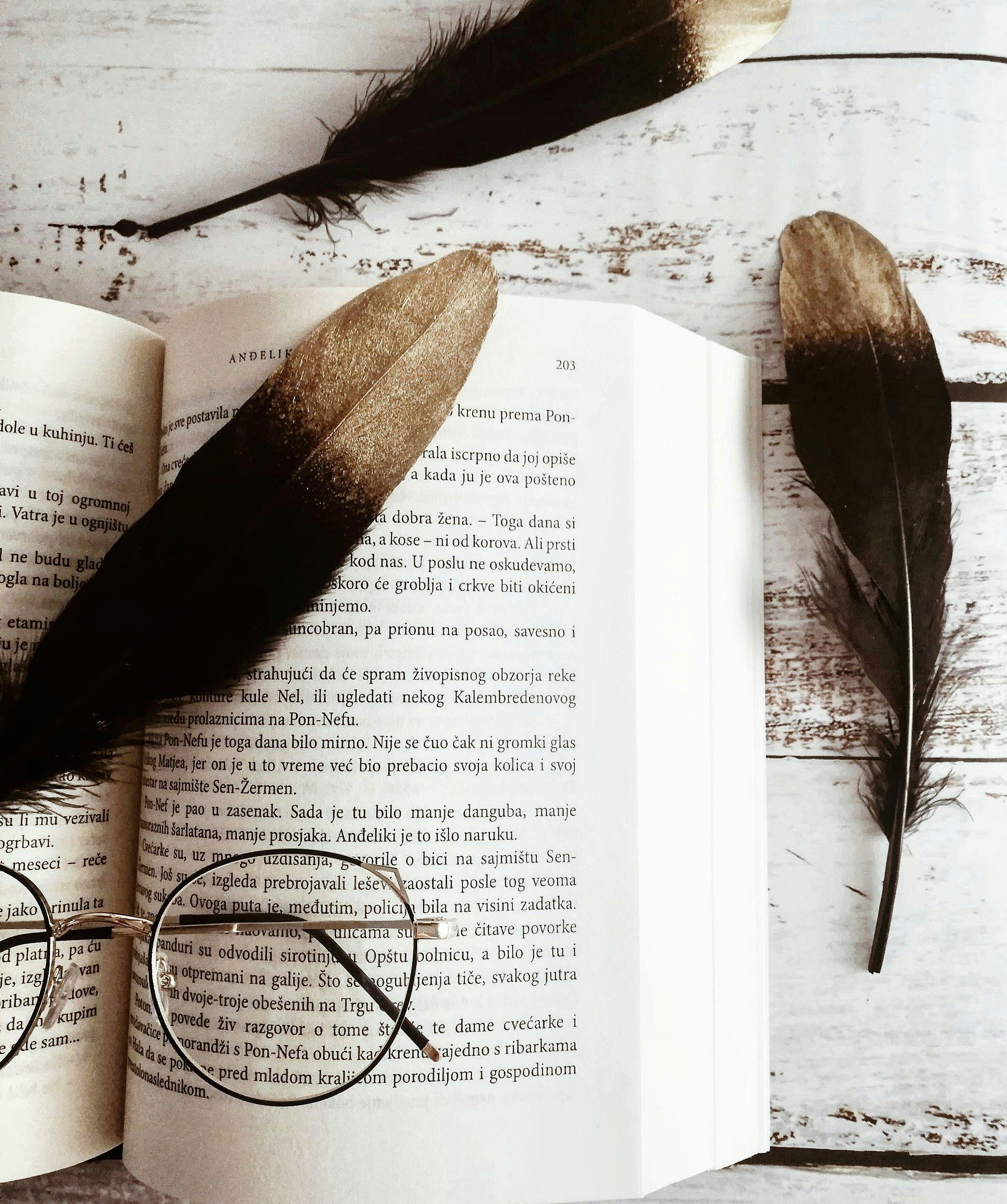 Un livre ouvert posé sur une surface en bois blanc, entouré de trois plumes noires et de lunettes rondes à monture métallique.