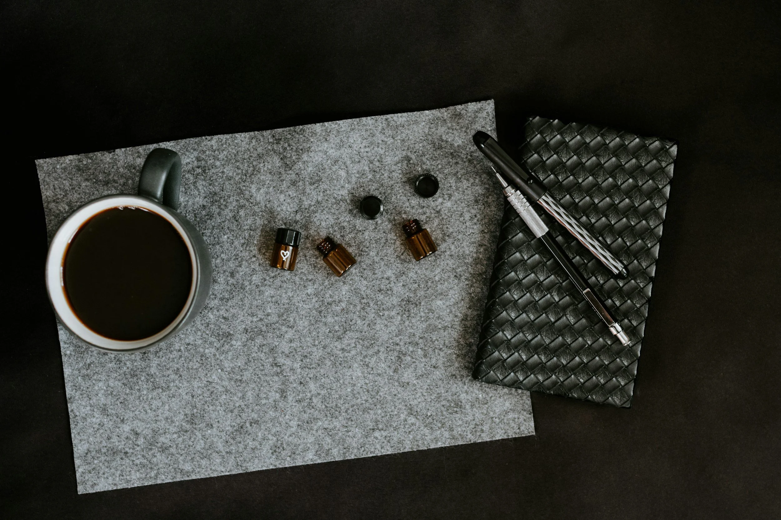 Une tasse de café noir, trois petites bouteilles d'huiles essentielles ou de médicaments, deux bouchons, un stylo et un porte-documents en cuir noir sur une surface grise, avec une feuille de papier.