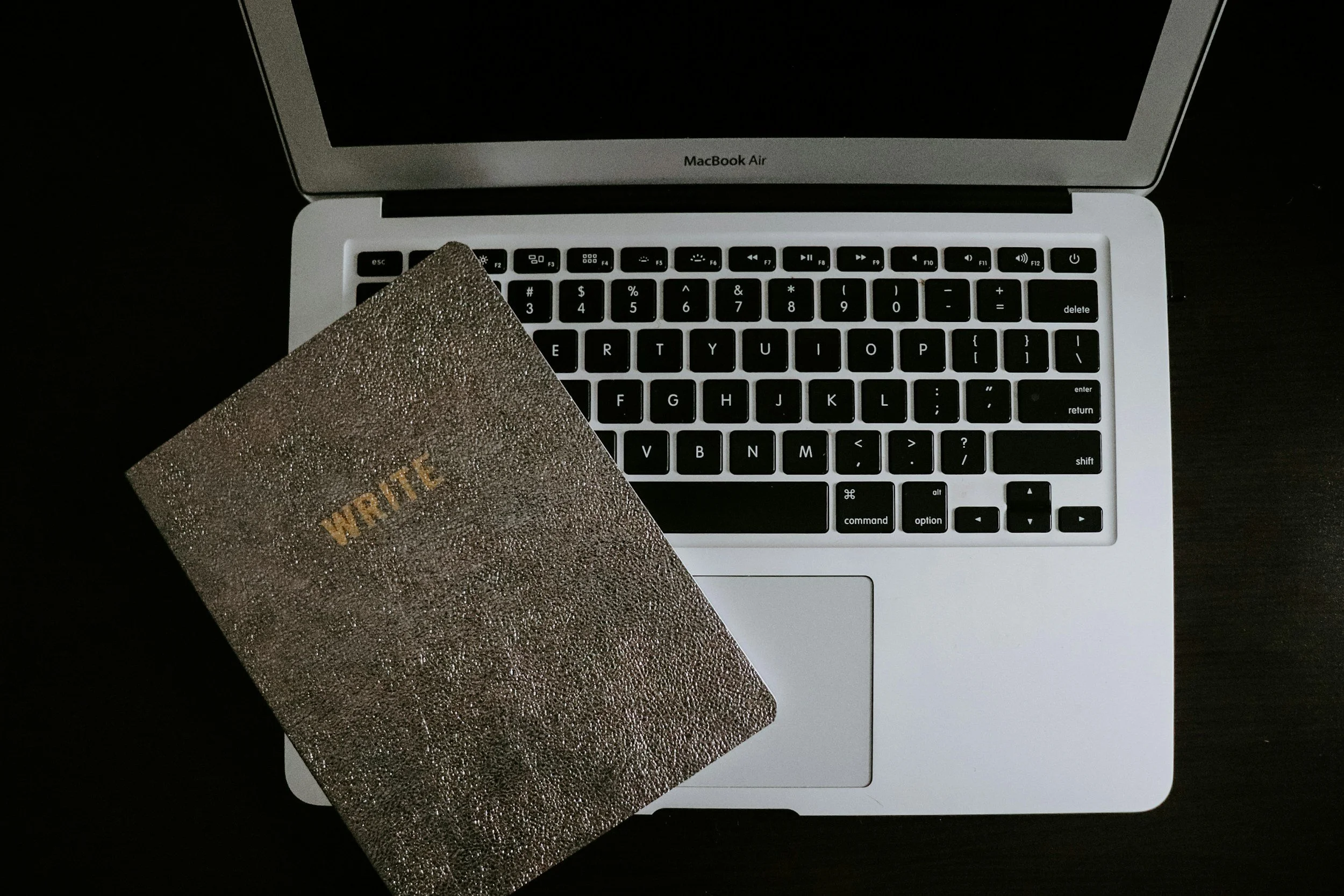 Un ordinateur portable MacBook Air ouvert avec un clavier noir et blanc, un écran noir, et un cahier ou un agenda orné de pierres brillantes, avec le mot 'WAIT' écrit en or