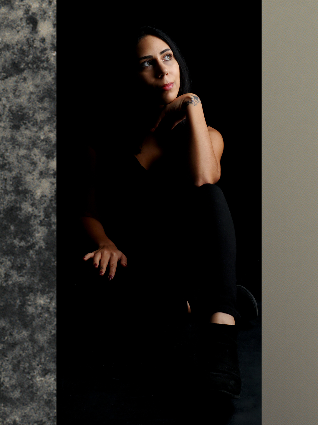 Femme aux cheveux noirs, regardant vers la droite, assise dans l'ombre, éclairée partiellement, portant des vêtements noirs.