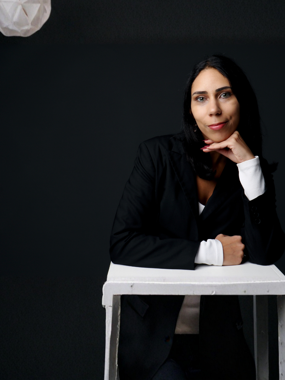 Portrait d'une femme avec des cheveux noirs, portant un blazer noir avec des manches blanches, assise à une table blanche contre un fond noir.