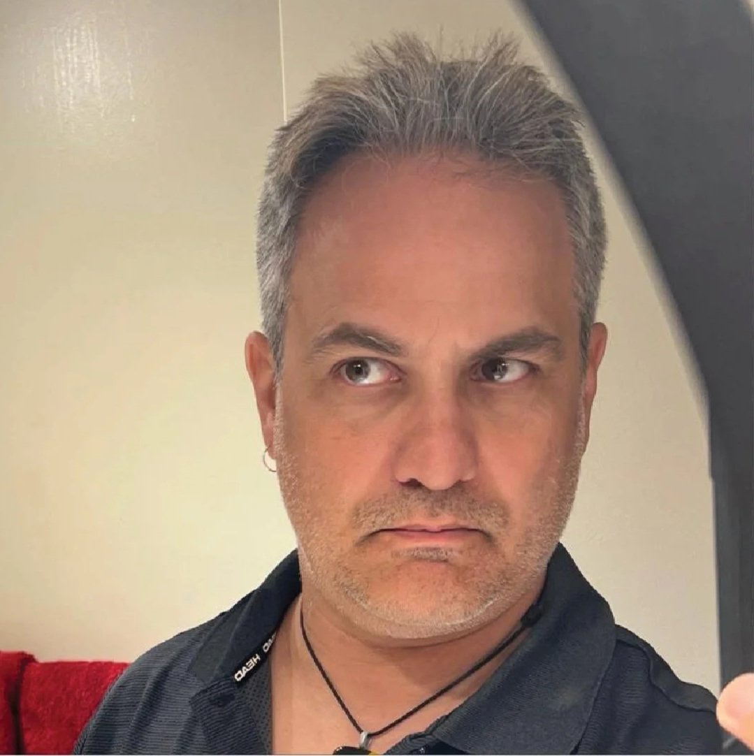 A middle-aged man with salt-and-pepper hair taking a selfie in front of a mirror, wearing a black shirt and a necklace.