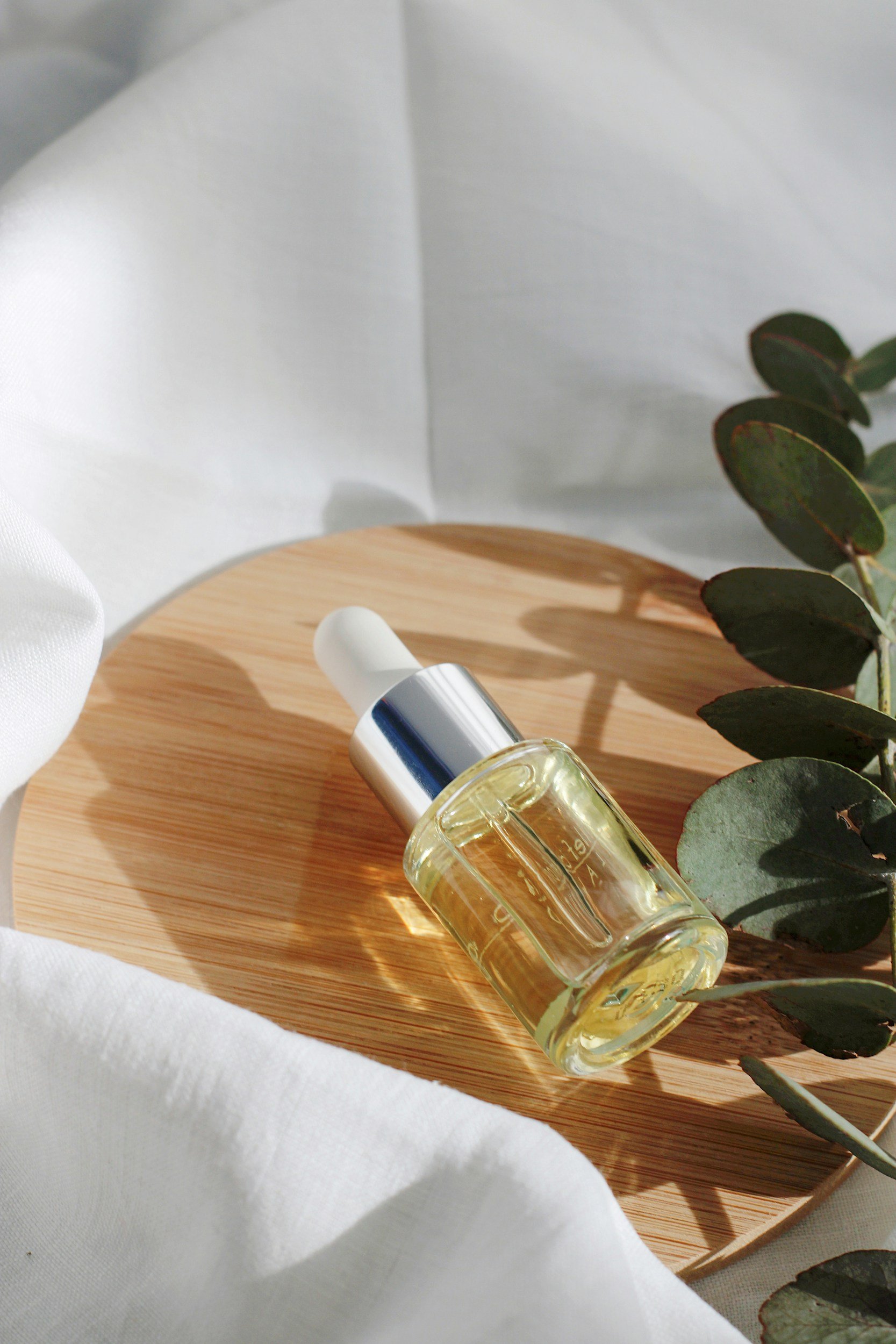 A glass bottle of facial oil with a dropper cap on a wooden plate, with eucalyptus leaves and white fabric in the background.