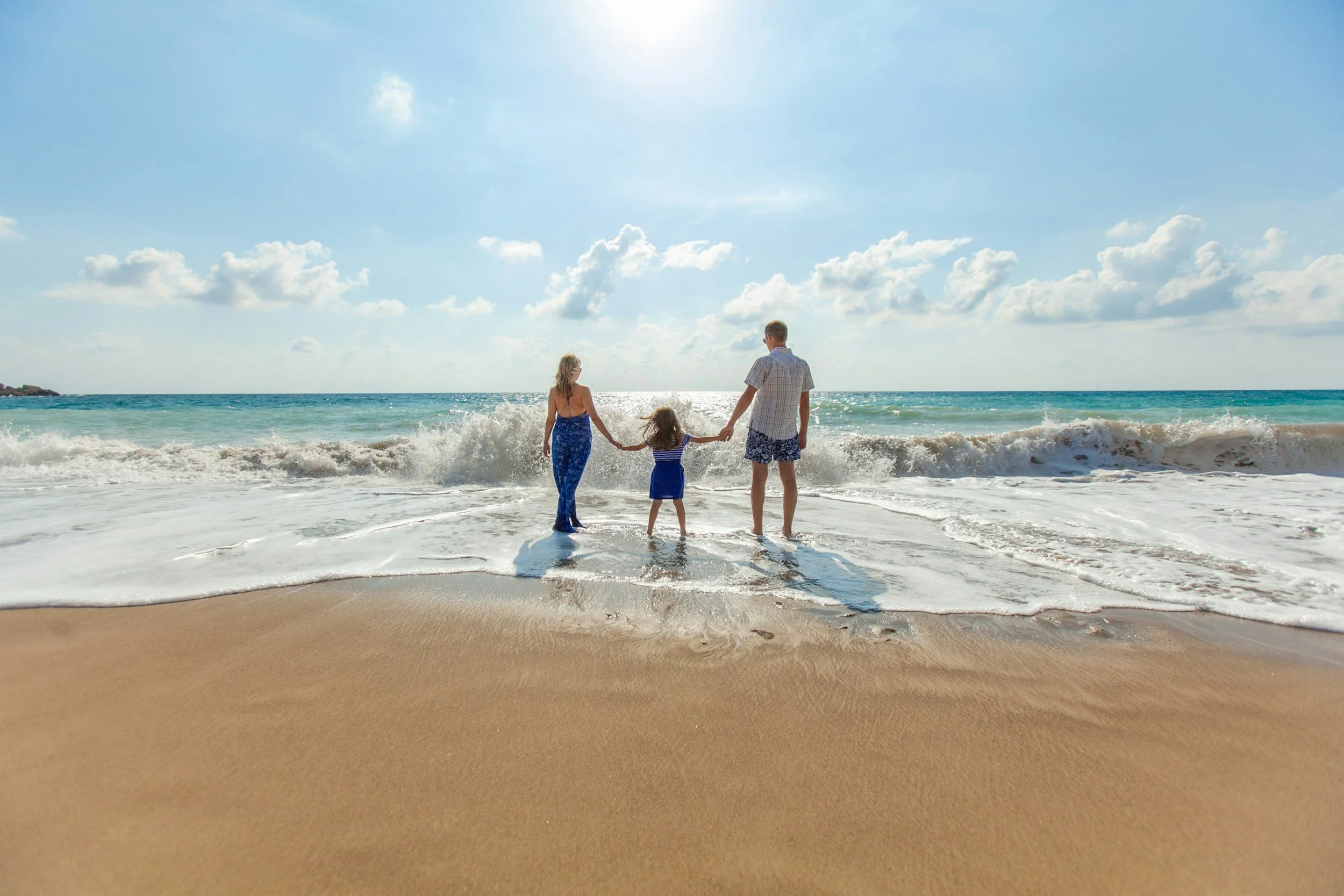 A family of three holding hands and standing at the edge of the ocean on a sunny beach.