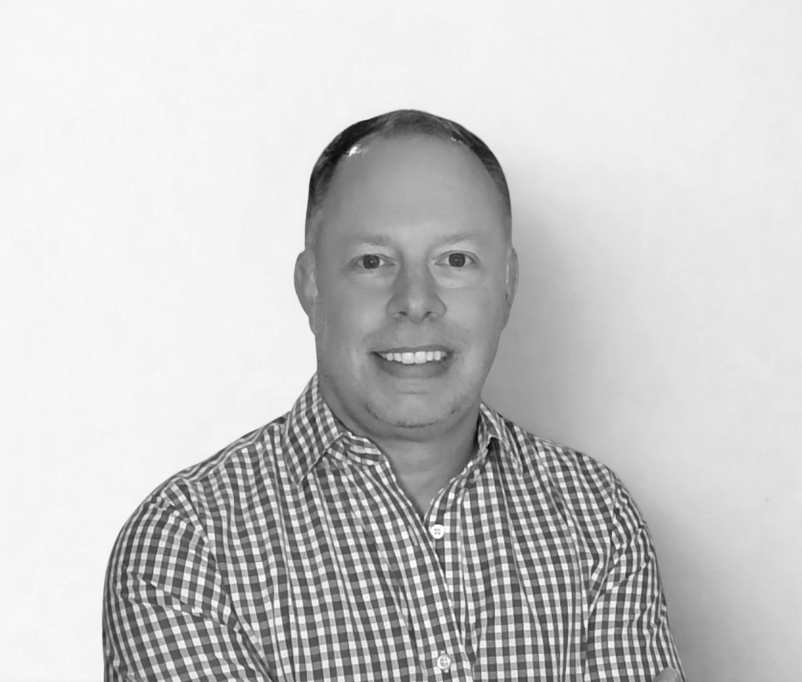 Black and white photo of a smiling man with short hair, wearing a checkered collared shirt, against a plain light background.