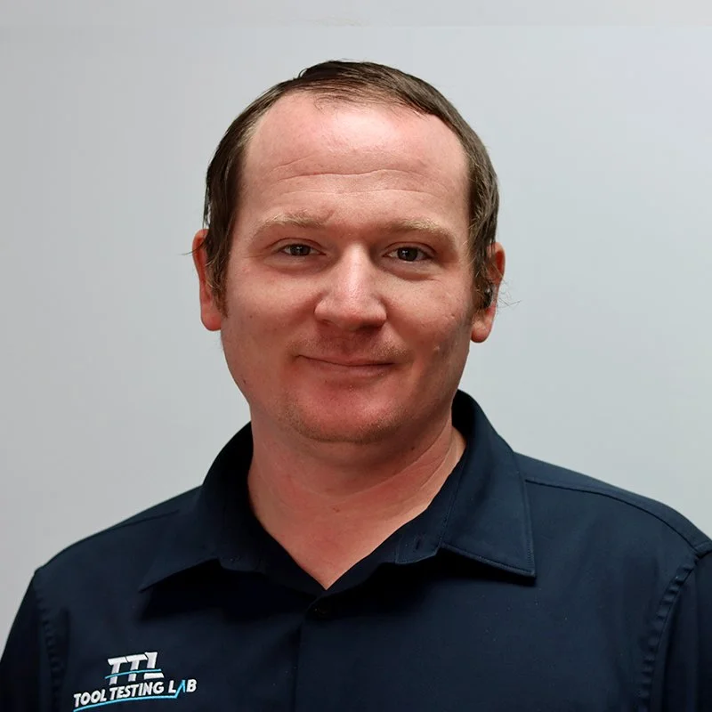 A man with glasses, a beard, and a bald head smiling, wearing a blue suit, light blue shirt, and red striped tie, standing outdoors against a gray textured wall.