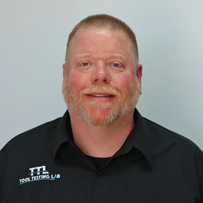 A man with glasses and a beard wearing a blue suit, light blue shirt, and a patterned tie, smiling in front of a concrete wall.