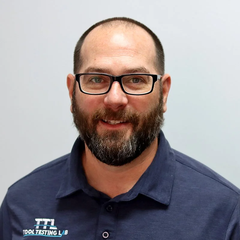 Portrait of a man with glasses, a beard, wearing a suit and tie, standing outdoors against a textured gray wall.