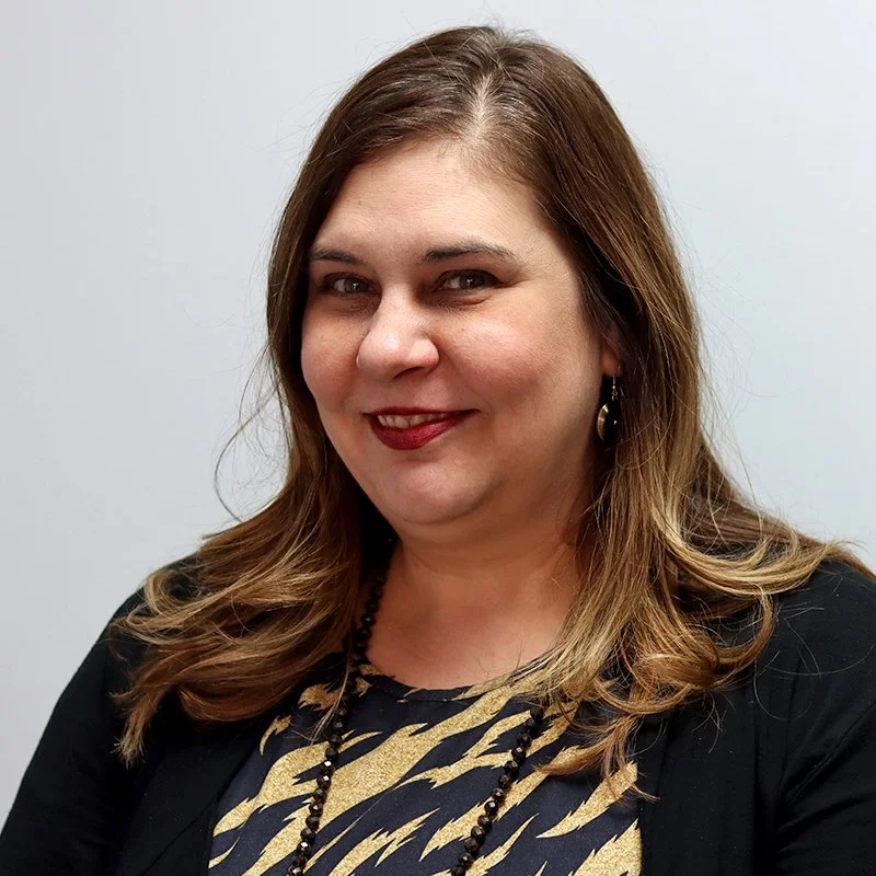 A man with glasses, a beard, and a bald head, smiling, wearing a blue suit and a checkered tie, standing in front of a concrete wall.