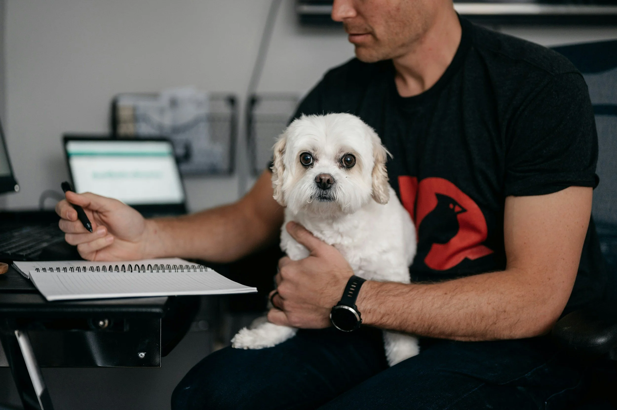 A man sitting at a desk holding a small white dog with light brown ears on his lap. The man is wearing a black T-shirt with a red graphic, a black watch, and a black wristband. On the desk are a spiral notebook, a laptop, and a pen. In the background, there are office shelves with storage boxes.