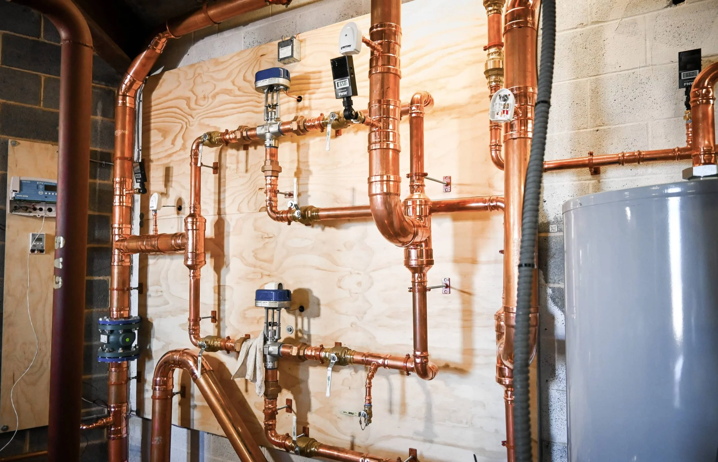 Copper plumbing pipes and gauges mounted on a wooden board in a utility room.