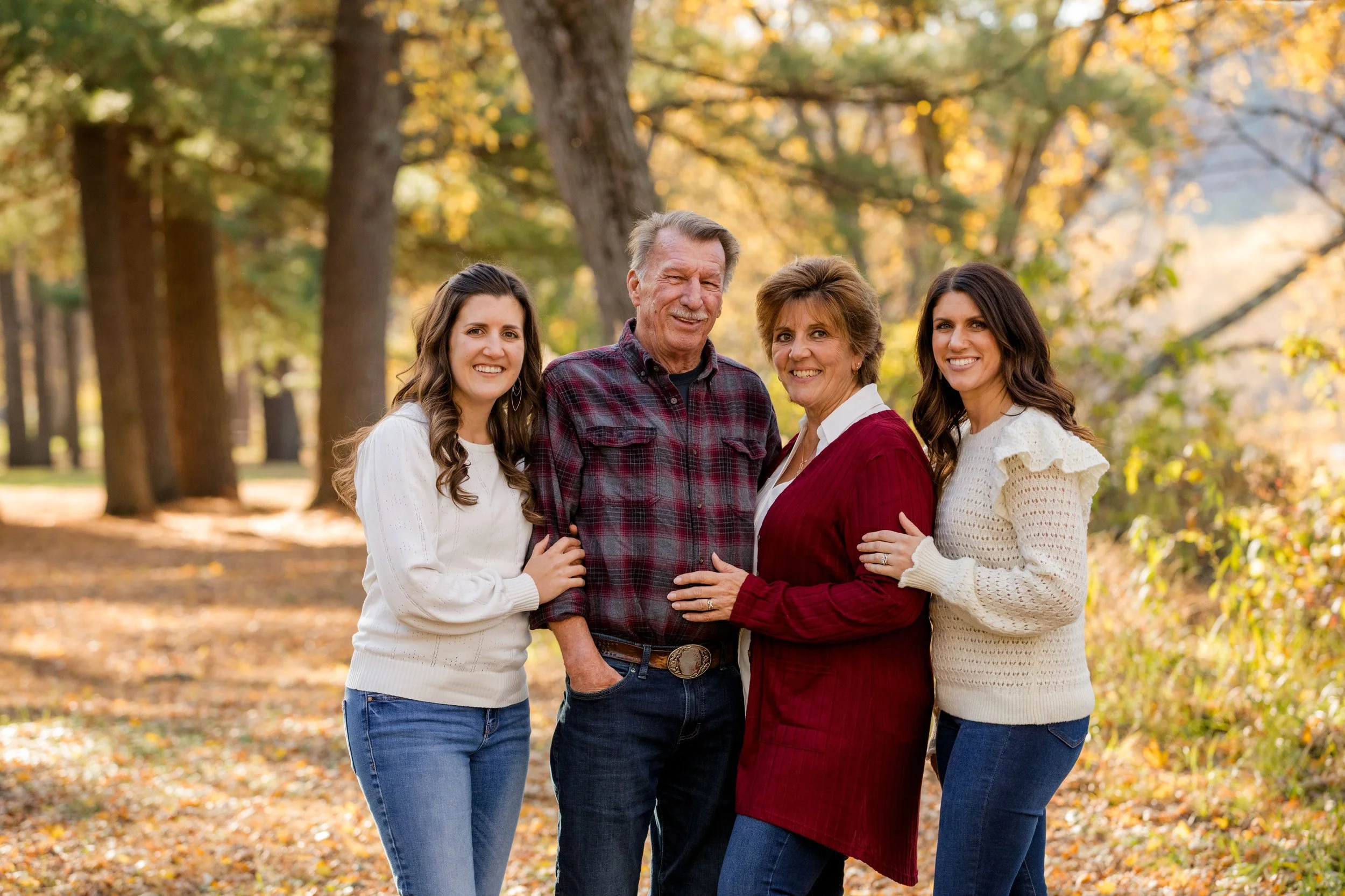 Family of four posing outdoors on a fall day with trees and colorful leaves in the background.