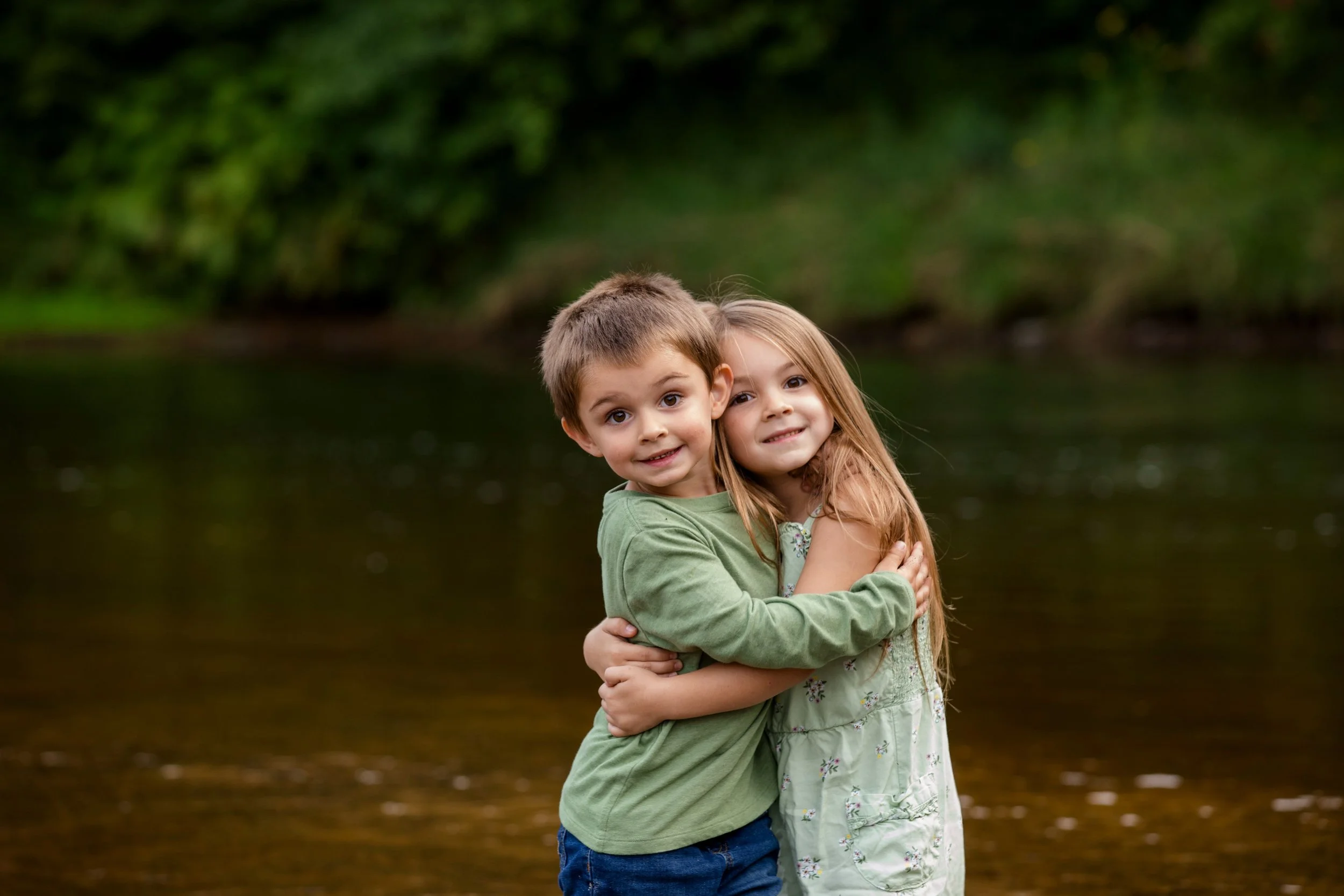 A young boy and girl hugging by a river with trees in the background.