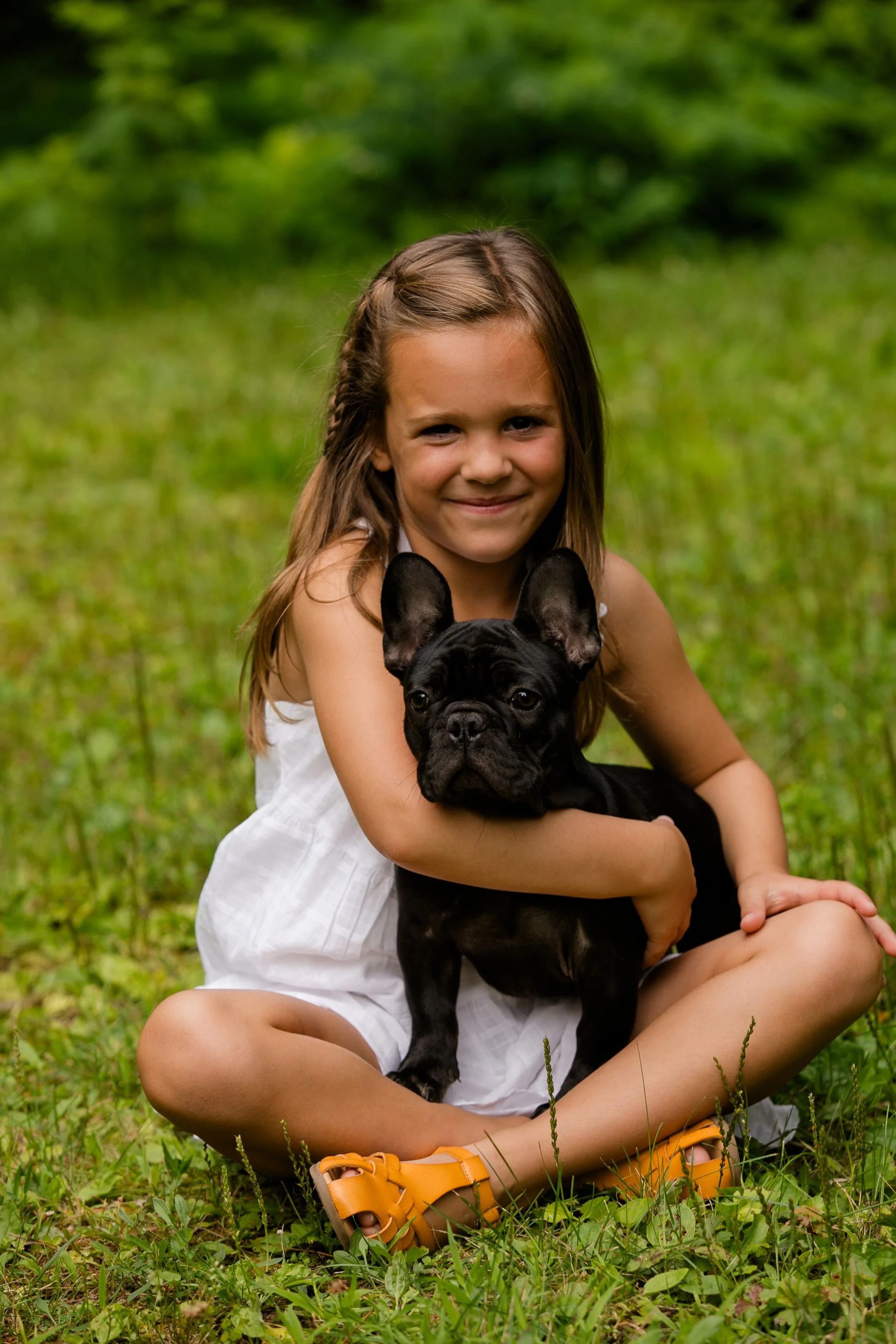 A young girl sitting cross-legged on the grass, smiling while holding a black French Bulldog puppy.