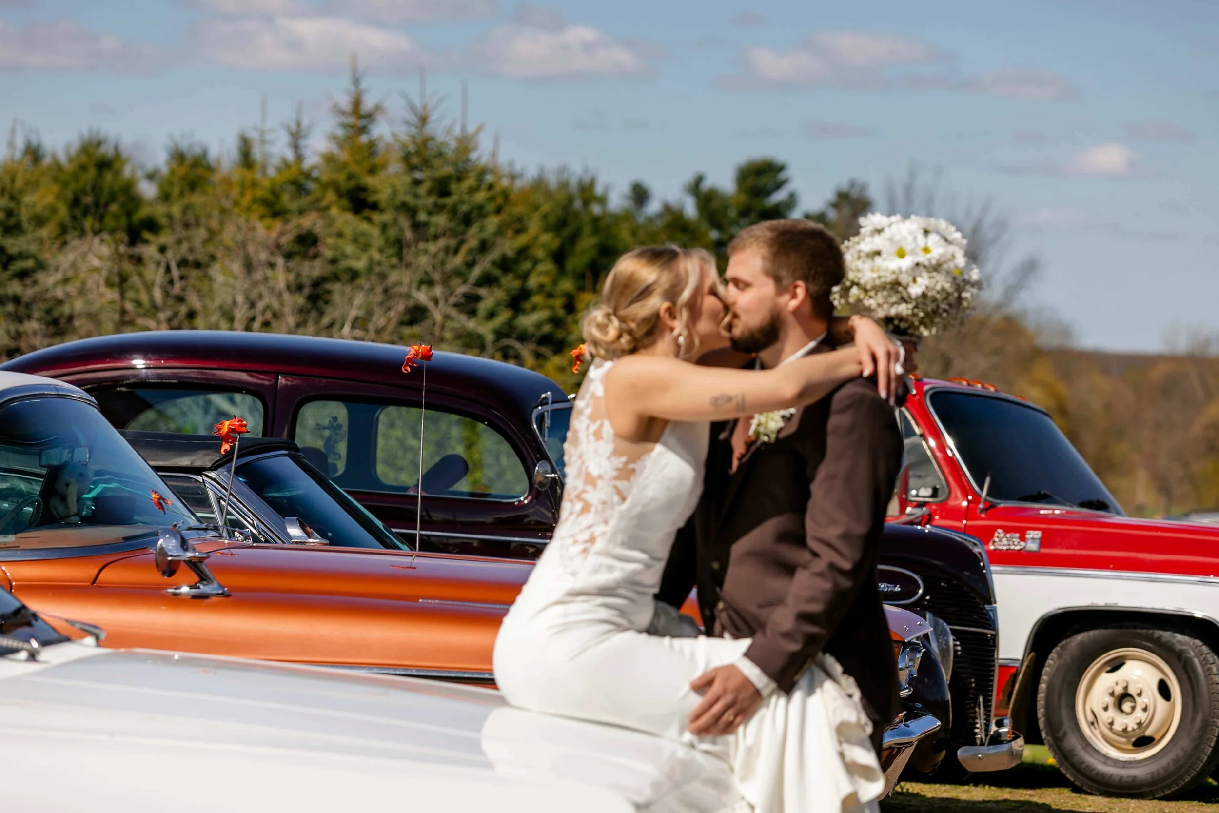 bride sitting on antique car with her arms around his neck and the groom kissing her on lips out of focus