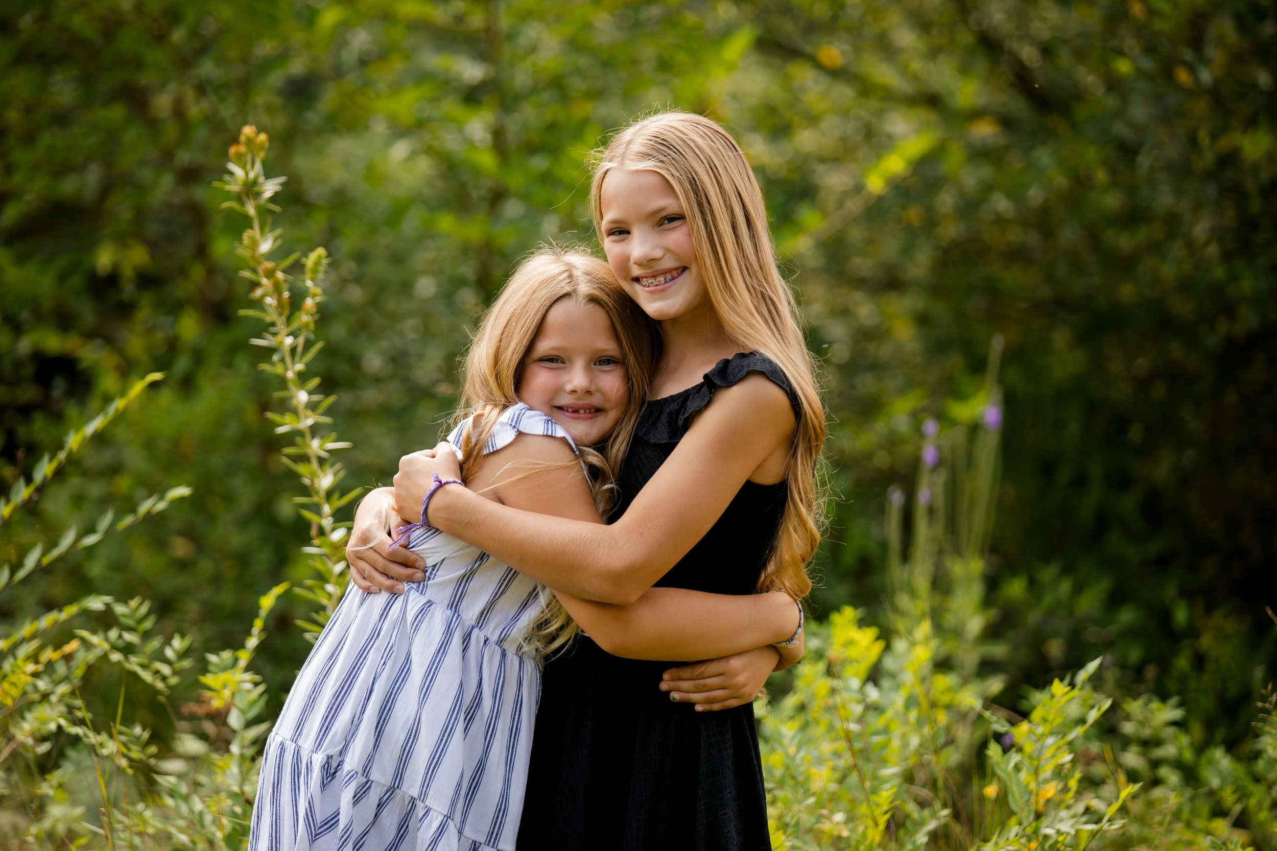 Two young girls with long hair, one in a black dress and the other in a white dress, hugging outdoors in a green, natural setting with bushes and trees in the background.