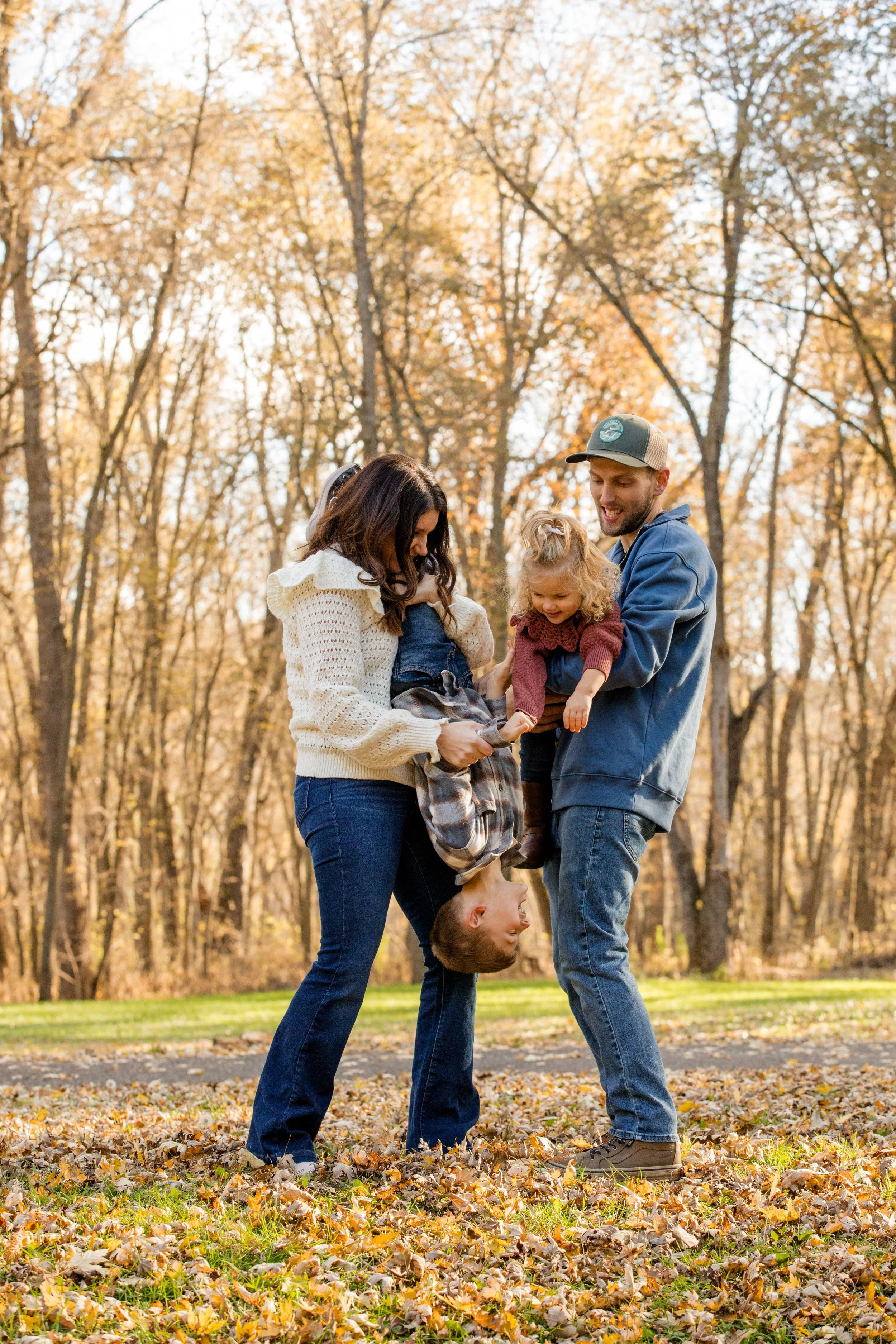 A family of four playing together in a park during fall. The mother and father are holding their two young children, one child upside down.