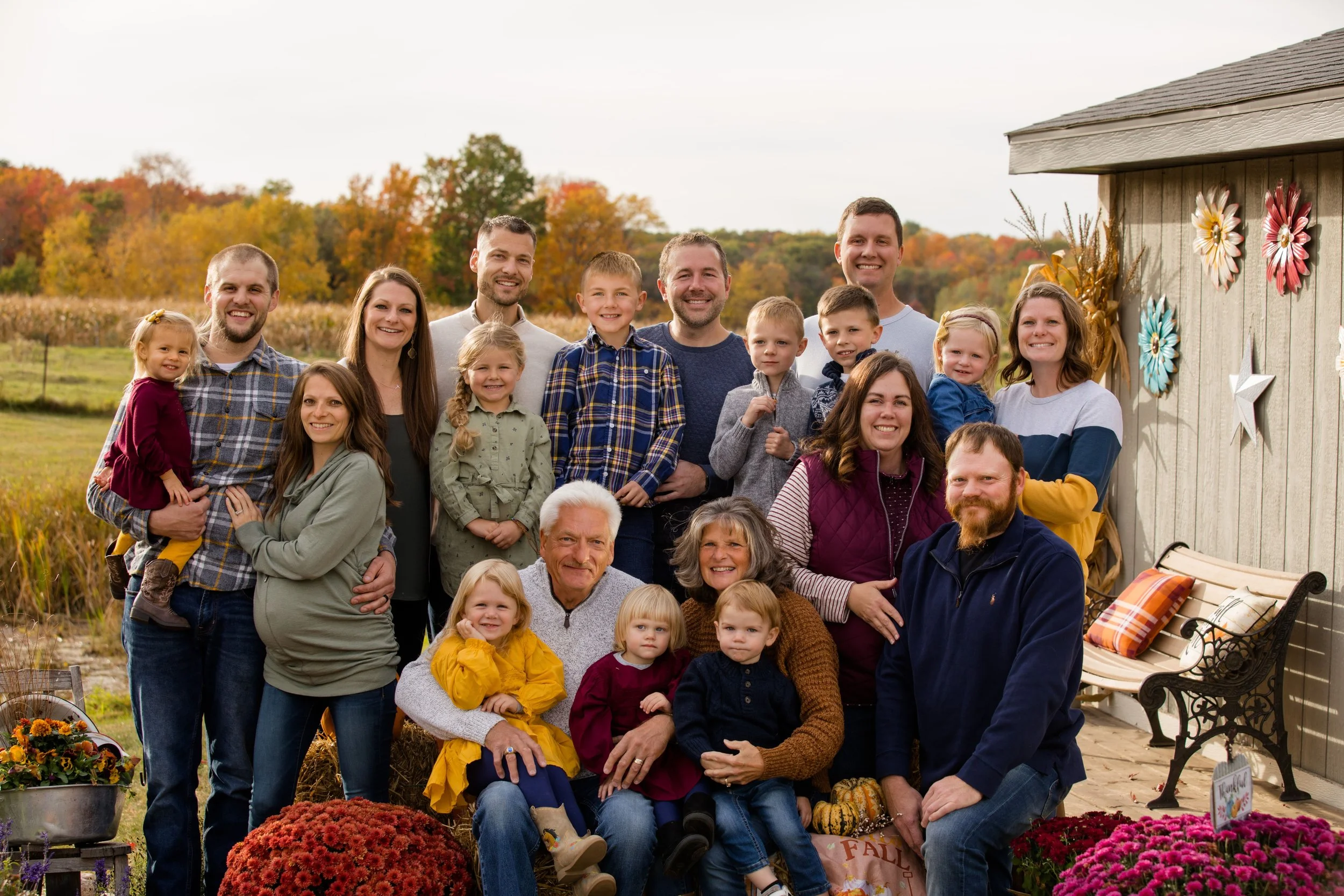 A large family photo outdoors in fall, with trees changing color in the background. The family members are smiling and gathered closely together, with some sitting and others standing. There is a rustic building with seasonal decorations and flower arrangements nearby.