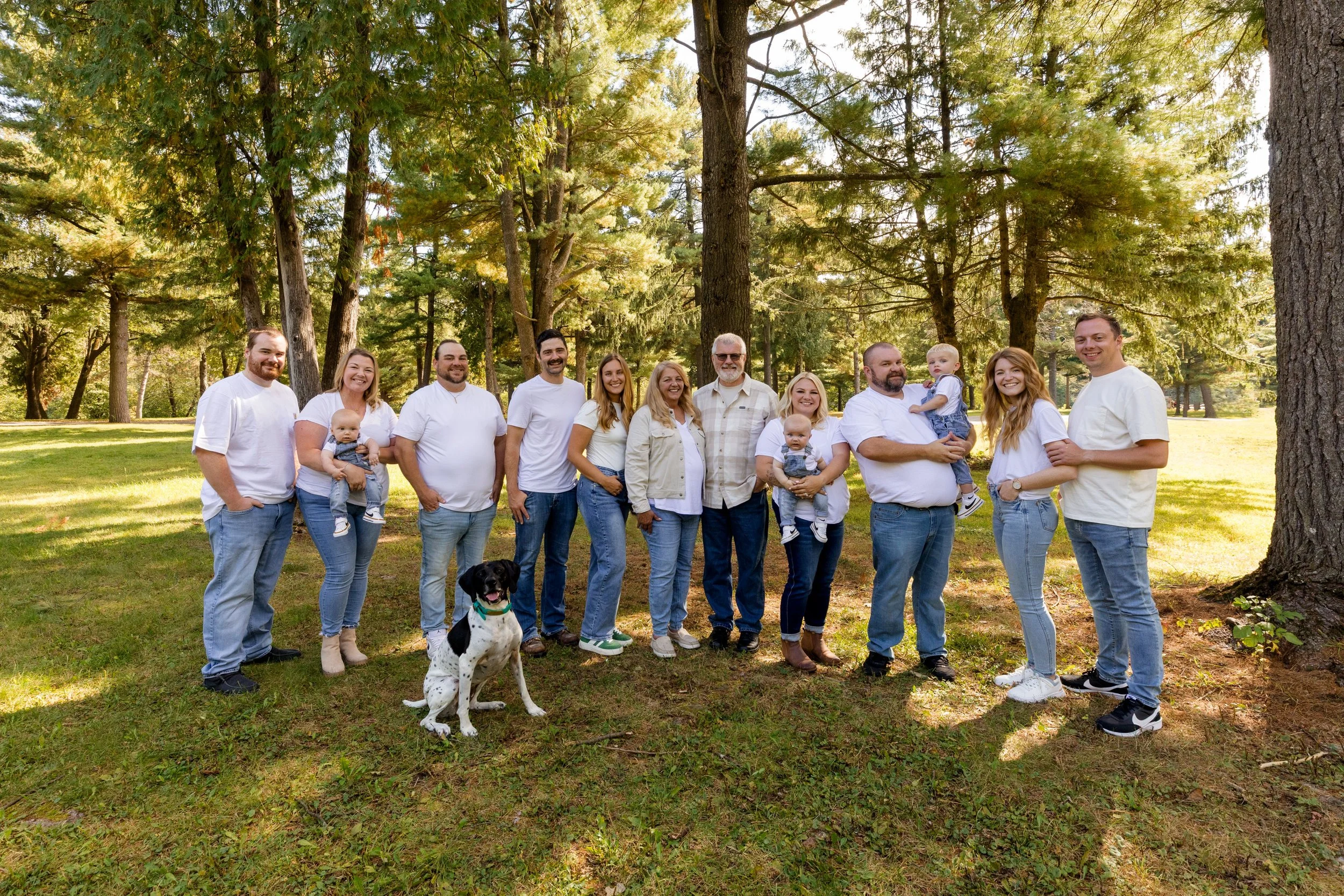 A large family gathered outdoors in a park with tall trees, posing for a group photo, with two small children, a dog, and everyone smiling.
