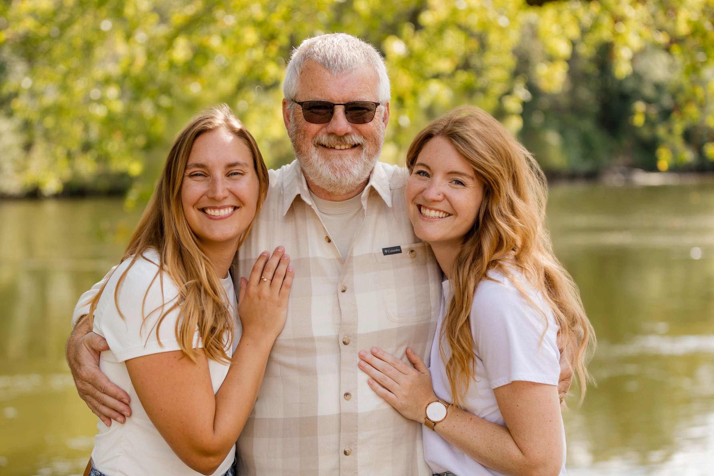 Three people smiling and hugging outdoors near a body of water, with trees in the background.