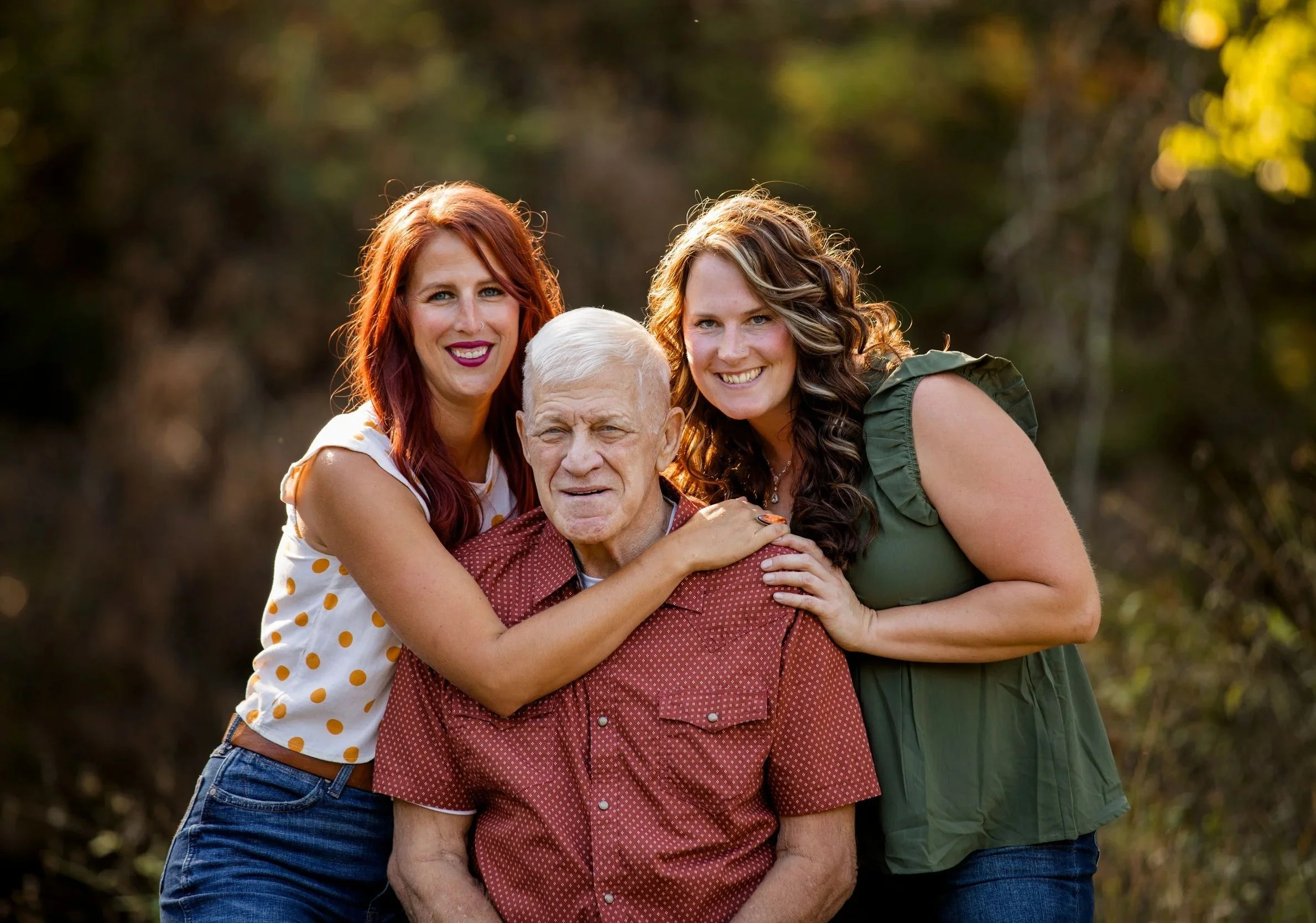 Two women and an elderly man smiling outdoors with a blurred green and yellow background. The women are standing close, with their arms around the man, who is seated.