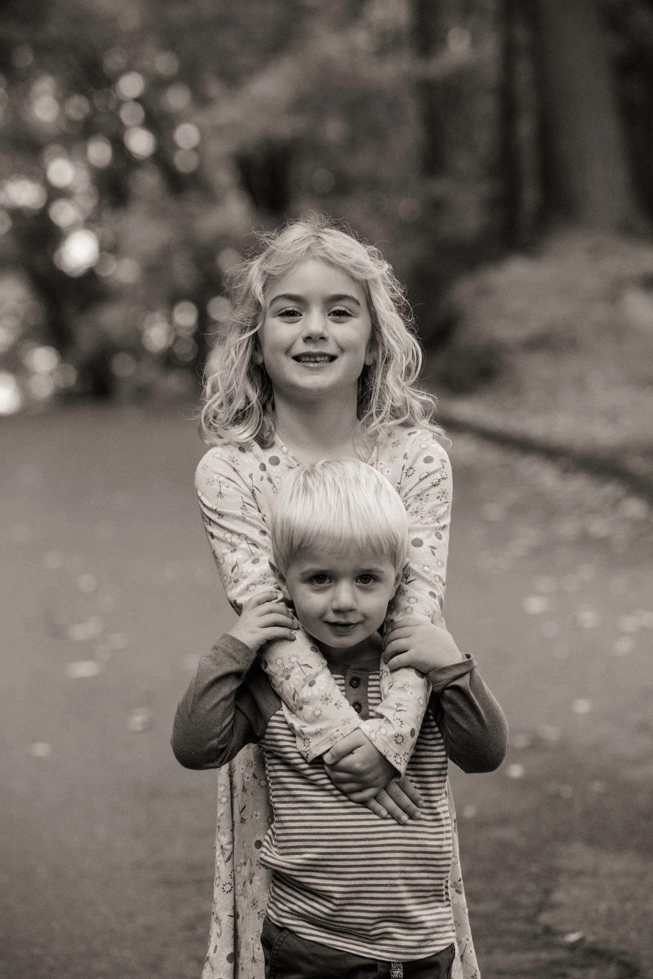 Two children, a girl with curly hair and a boy with straight hair, playing outdoors in a wooded area.