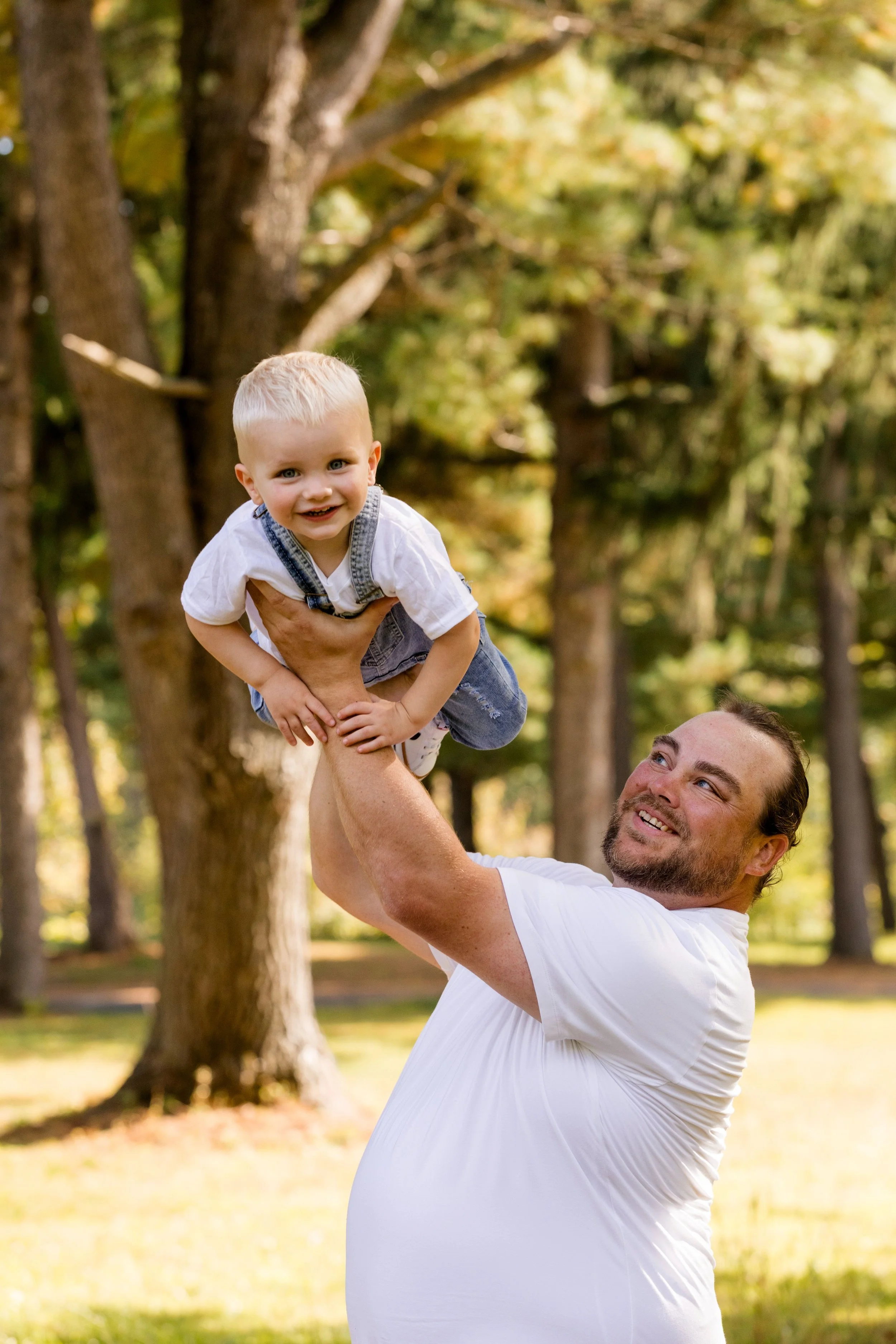 A man holding a young boy up in the air outdoors in a park with trees in the background.