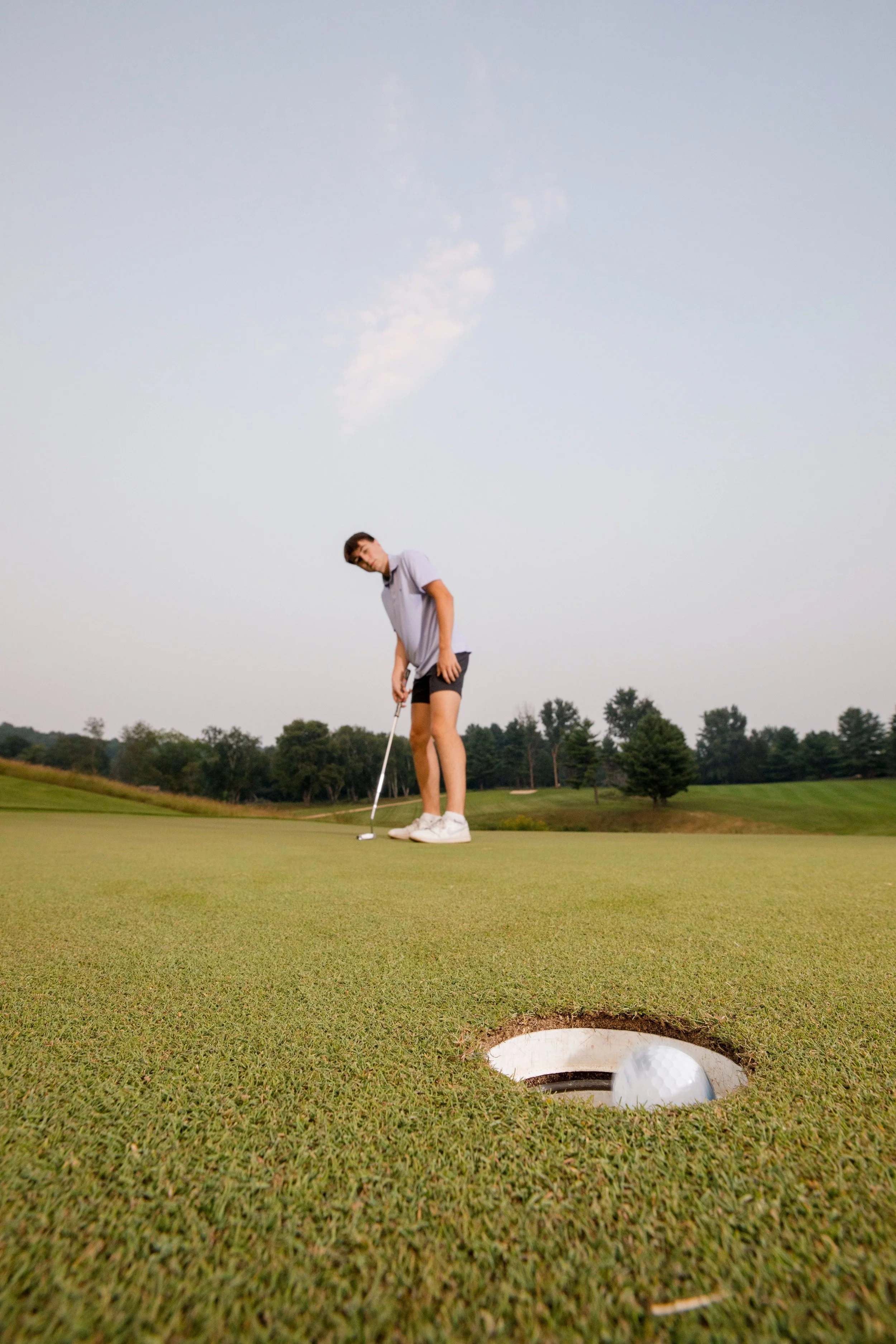 A young man preparing to putt a golf ball on a golf green outdoors, with a hole in the foreground and trees in the background for his high school senior pictures.