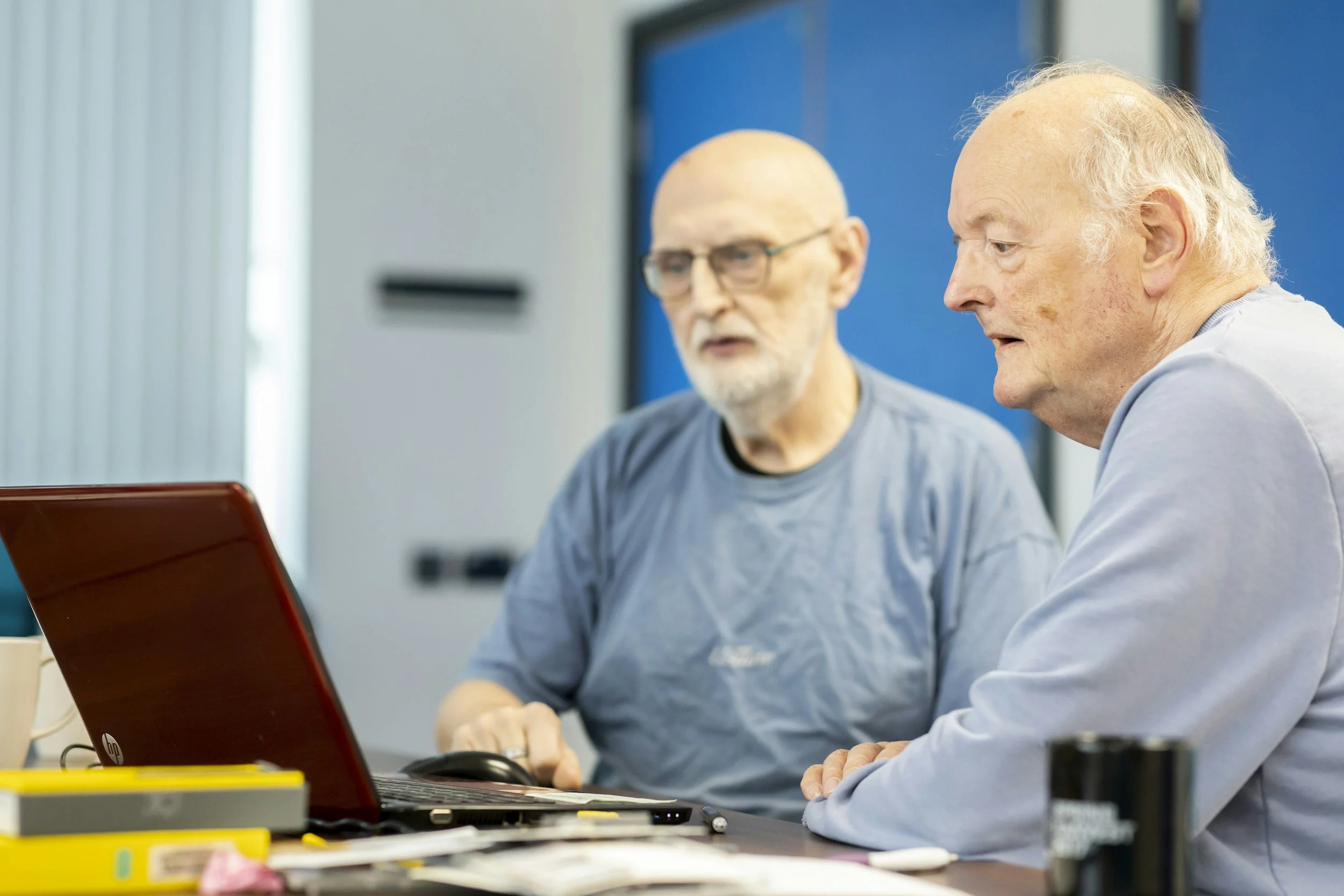 Two elderly men sitting at a desk looking at a laptop, in a bright room with blue and white walls.