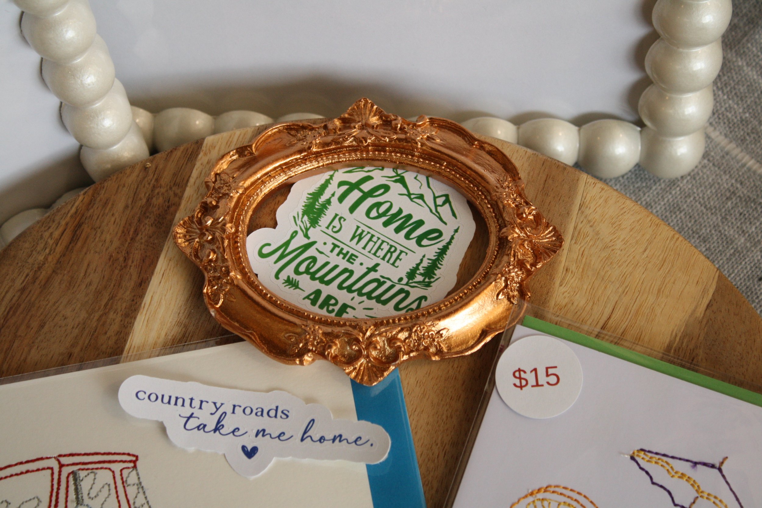 Close-up of a decorative copper-colored oval frame with green and white text, resting on a wooden surface. The text reads, 'Home is where the mountains are.' Part of a white beaded frame and a sign with the price '$15' are also visible.