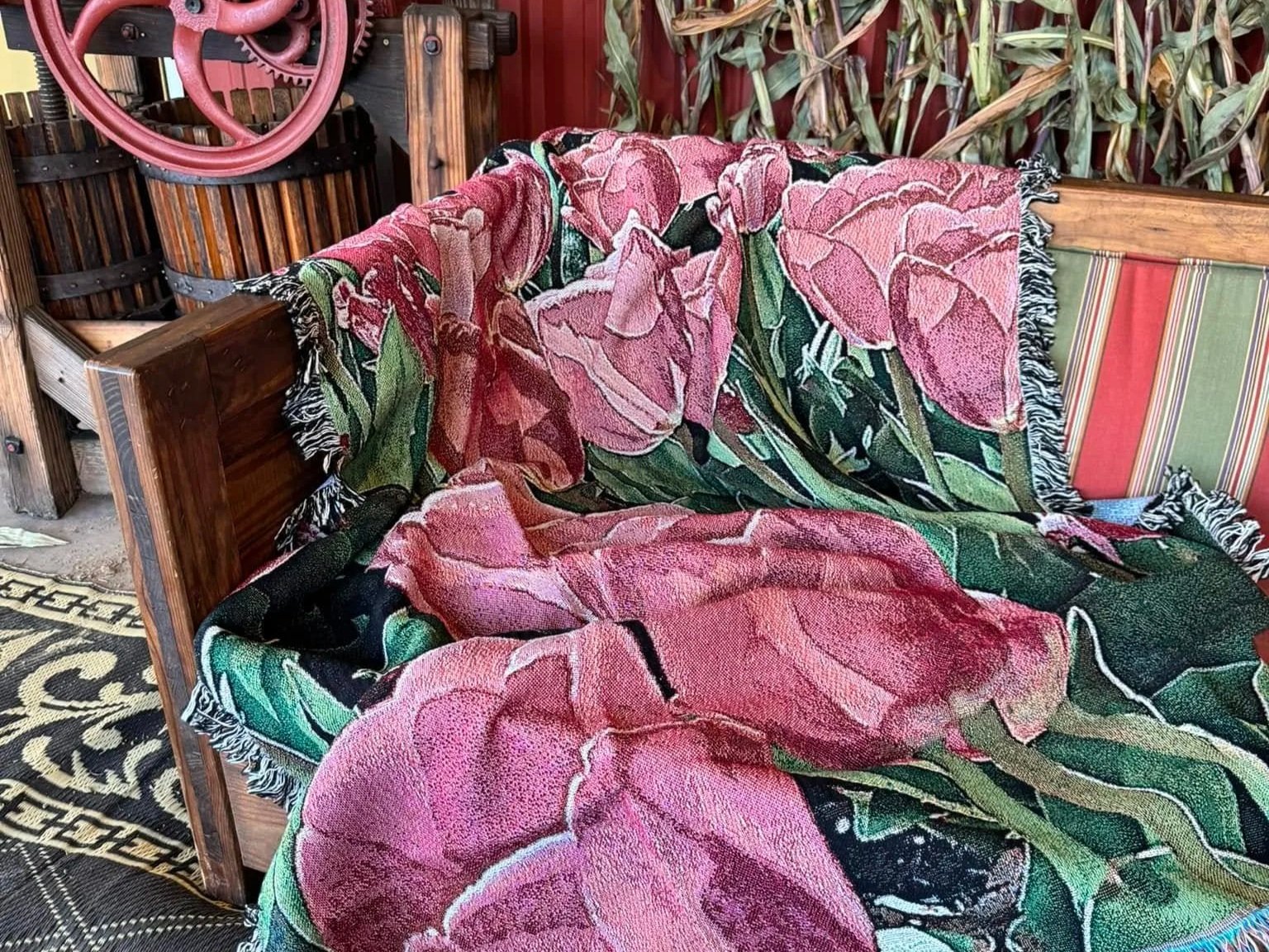 A floral-patterned blanket with pink and green colors draped over a wooden bench next to a striped cushion. In the background, there are dried plants or corn stalks and a piece of vintage farm equipment with a large red wheel.