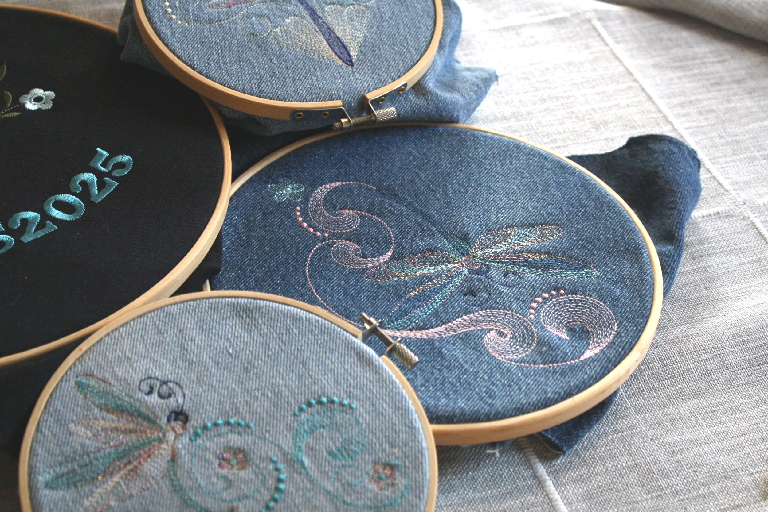 Close-up of several embroidery hoops with denim fabric, featuring designs of flowers, a dragonfly, and an abstract pattern.